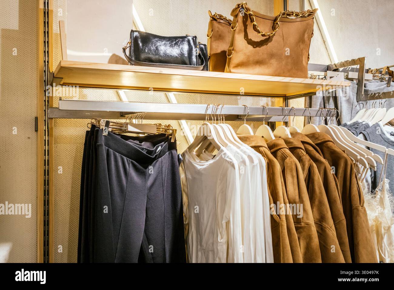 Un'elegante disposizione di abbigliamento da donna in un negozio, con camicette bianche, cappotti abbronzanti e pantaloni neri. Sopra gli indumenti, sono disponibili borse eleganti Foto Stock