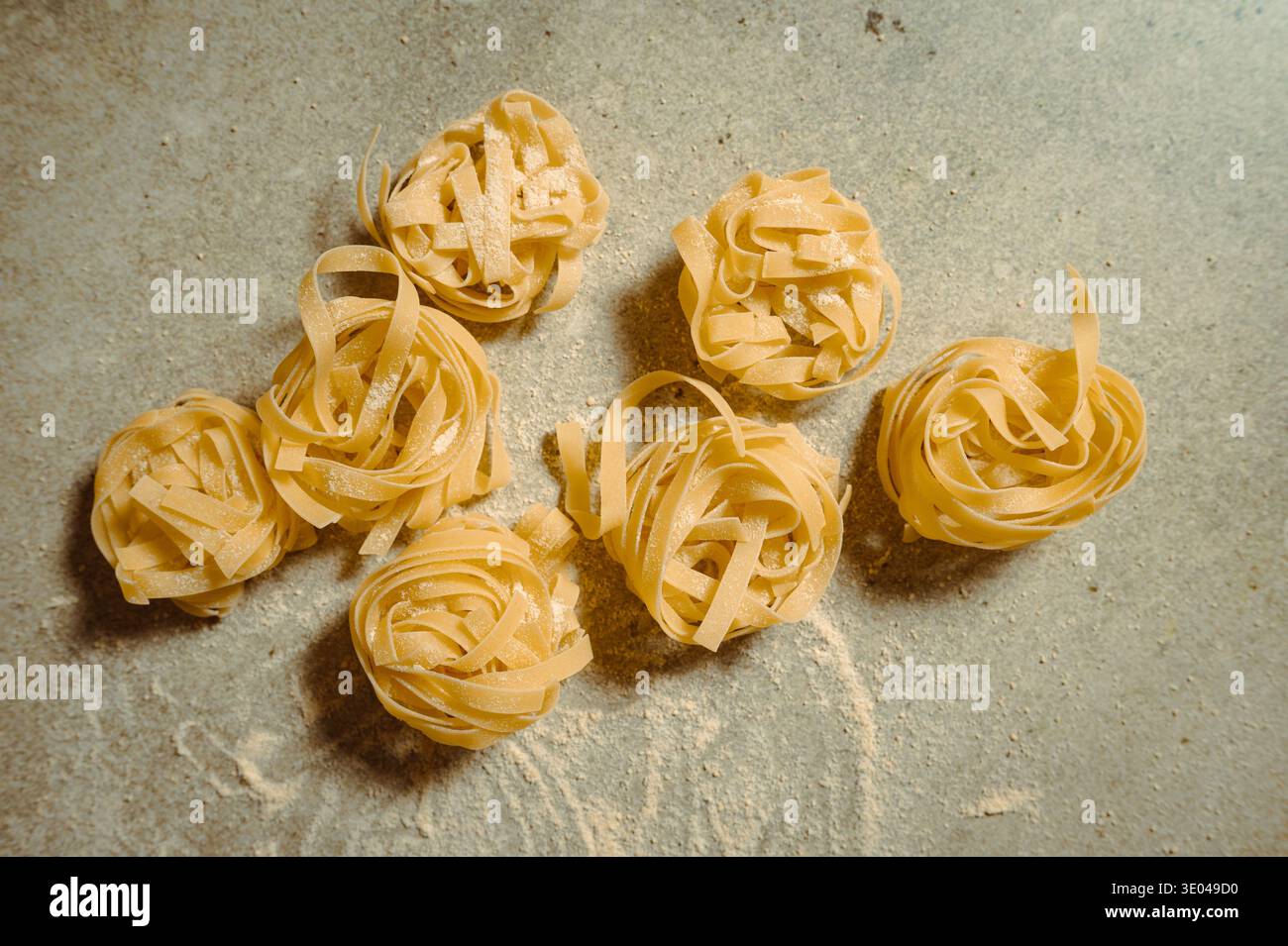 Vista dall'alto dei nidi di pasta appena fatti, disposti su una superficie infarinata. La vivace tonalità gialla della pasta cruda ne esalta la freschezza e la freschezza Foto Stock