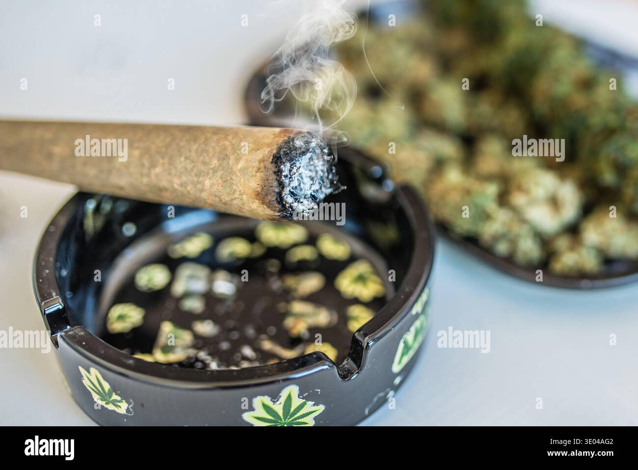 Primo piano di un giunto di cannabis illuminato che emette fumo accanto a un posacenere decorativo con disegni a foglia di cannabis, con gemme di cannabis sfocate sullo sfondo. Foto Stock