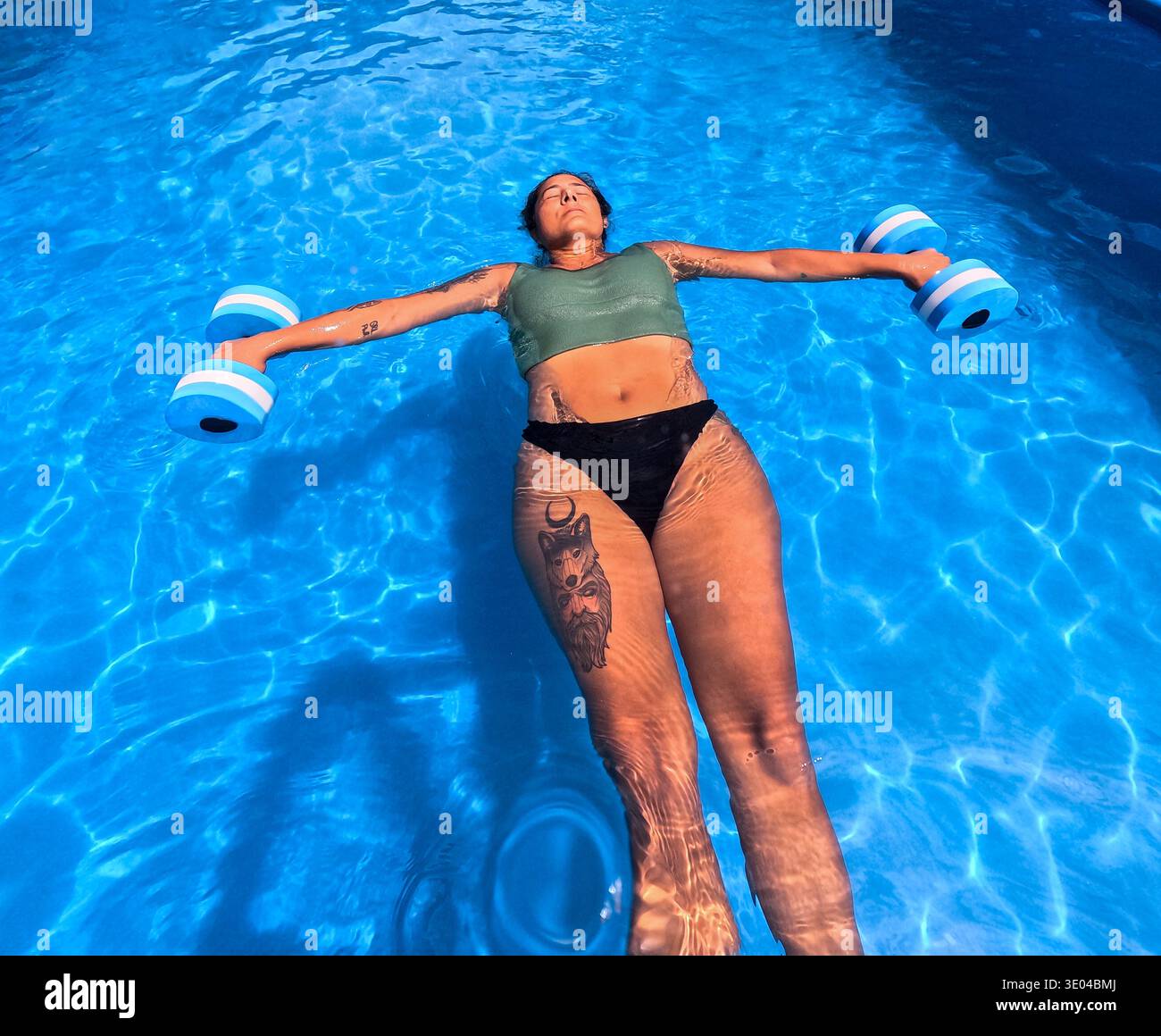 Una donna galleggia pacificamente in una piscina blu brillante, tenendo in mano pesi per un rilassante allenamento in acqua. La luce del sole crea splendidi riflessi Foto Stock