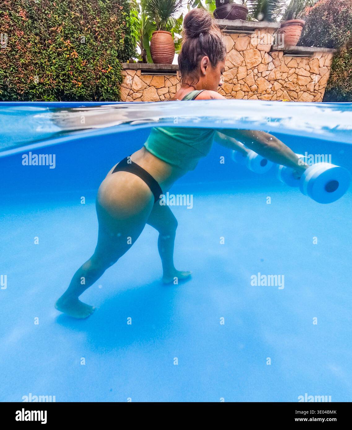 Donna impegnata nel fitness subacqueo usando pesi in acqua in una piscina, catturata in un ambiente vivace e chiaro. L'attività sottolinea la forza A. Foto Stock