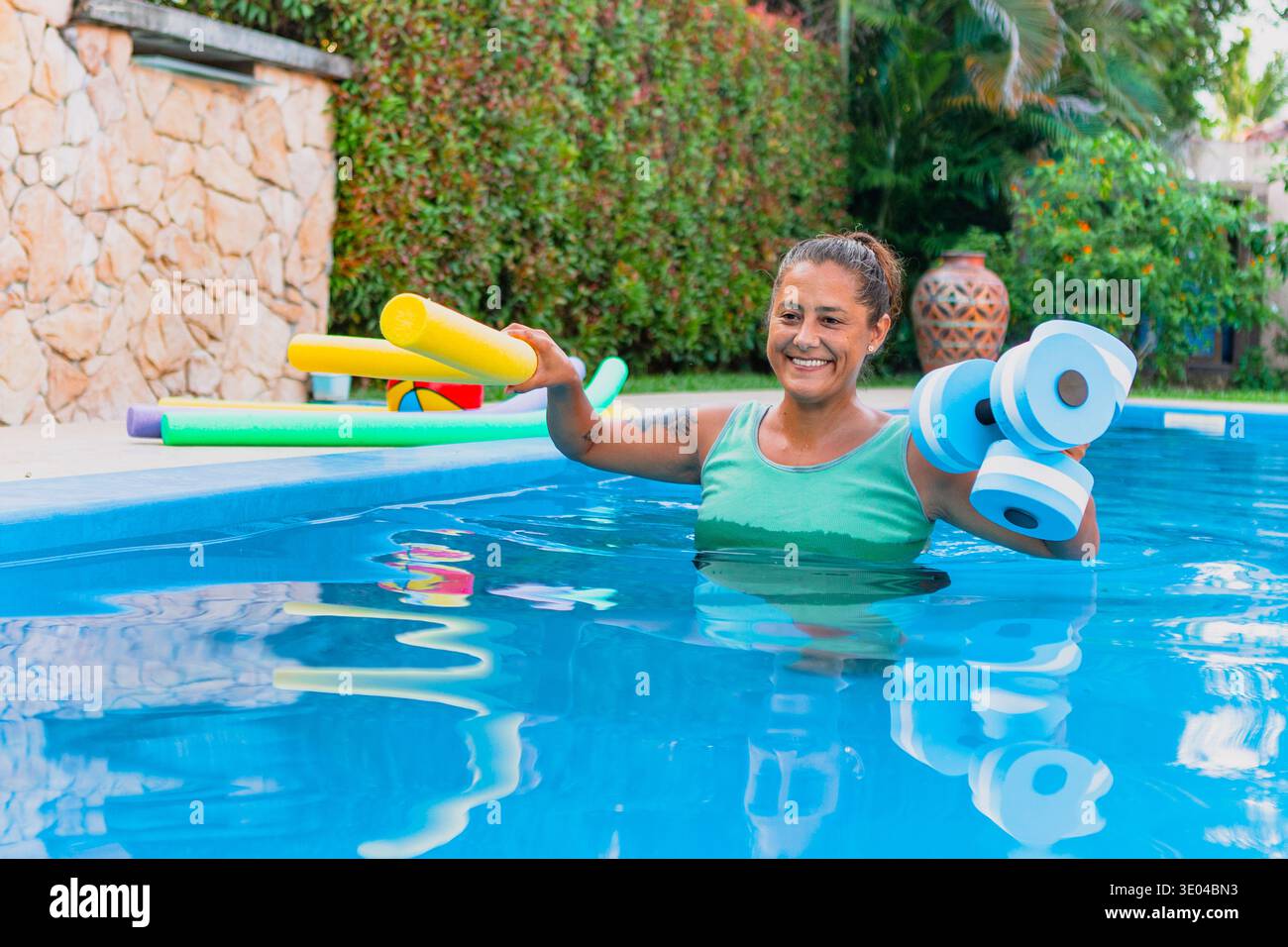 Una donna in piscina sorride mentre si fa esercizio con spaghetti e manubri in schiuma colorata. L'area a bordo piscina presenta una vegetazione lussureggiante e una pietra Foto Stock