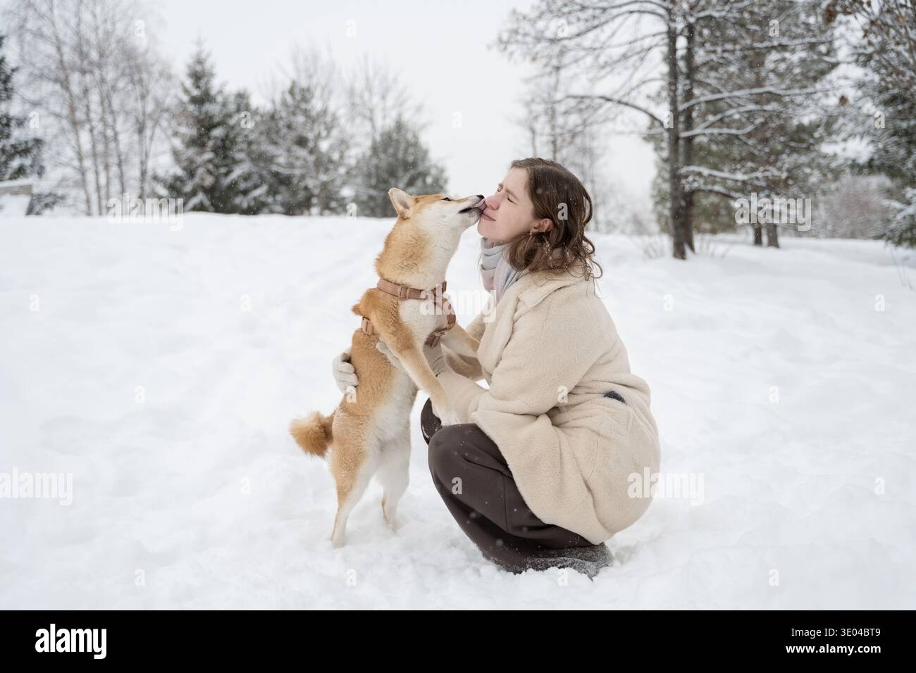Donna in camice beige inginocchiata nella neve mentre un cane si lecca amorevolmente il viso durante una passeggiata in un paese delle meraviglie invernale, circondato da alberi innevati. Foto Stock