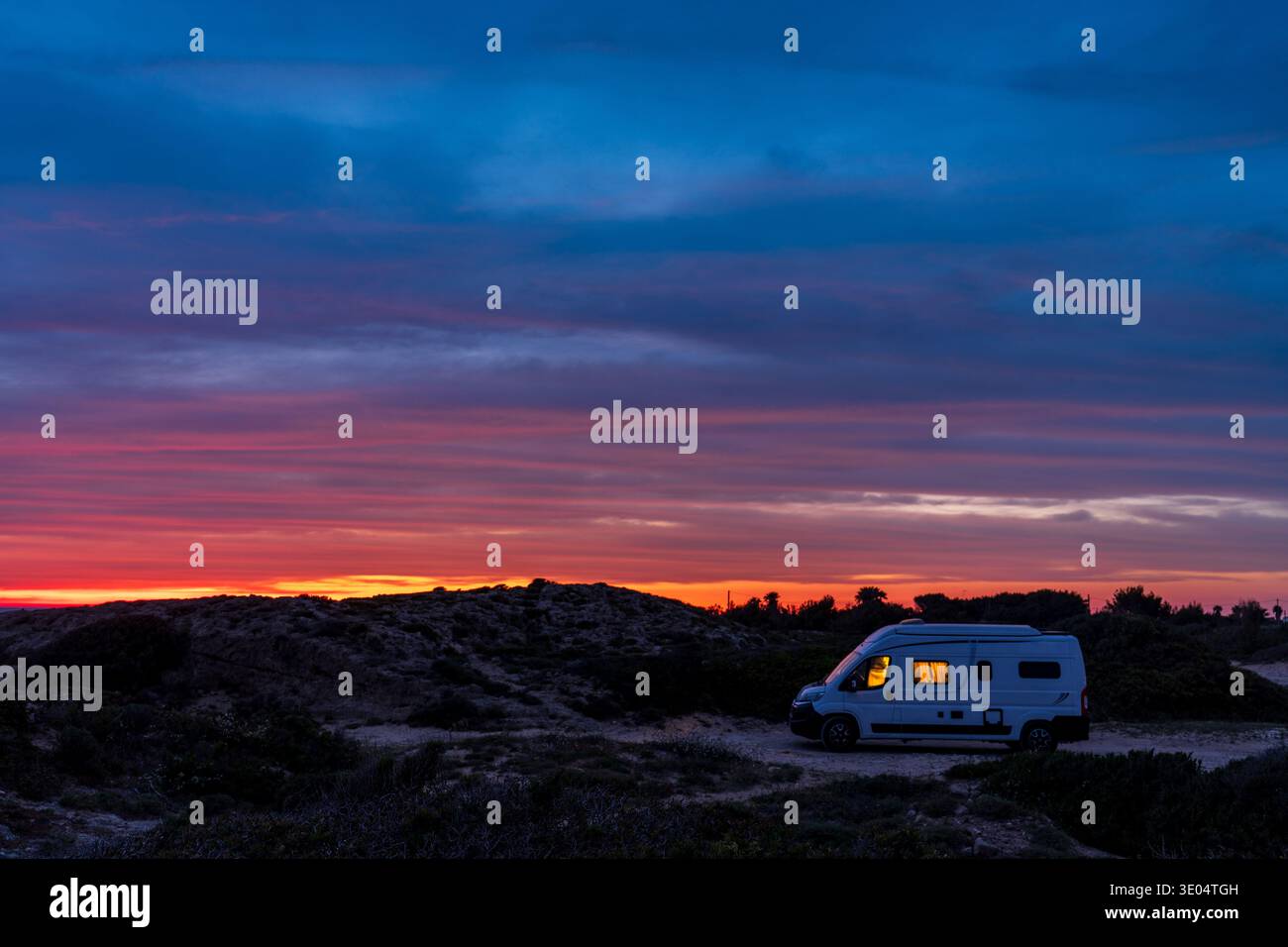 Un camper bianco parcheggiato tra le dune costiere al crepuscolo. Le finestre luminose emettono una luce calda in contrasto con un cielo spettacolare striato di arancione, Re Foto Stock