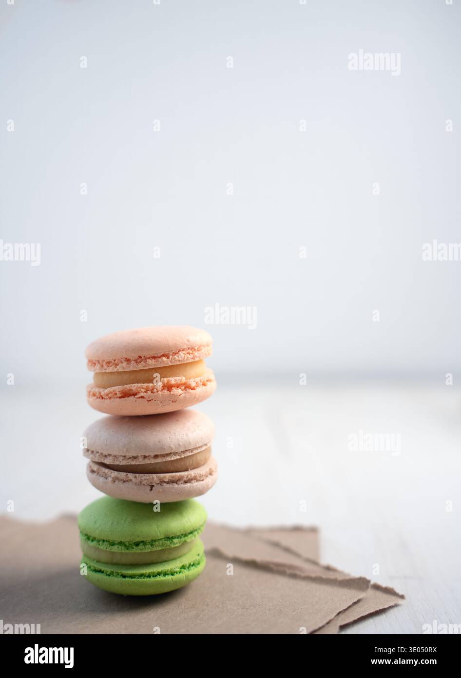 pile di tre macaron pastello (pesca, beige, verde) delicatamente bilanciati su carta kraft strappata su uno sfondo bianco brillante e minimalista da tavolo Foto Stock