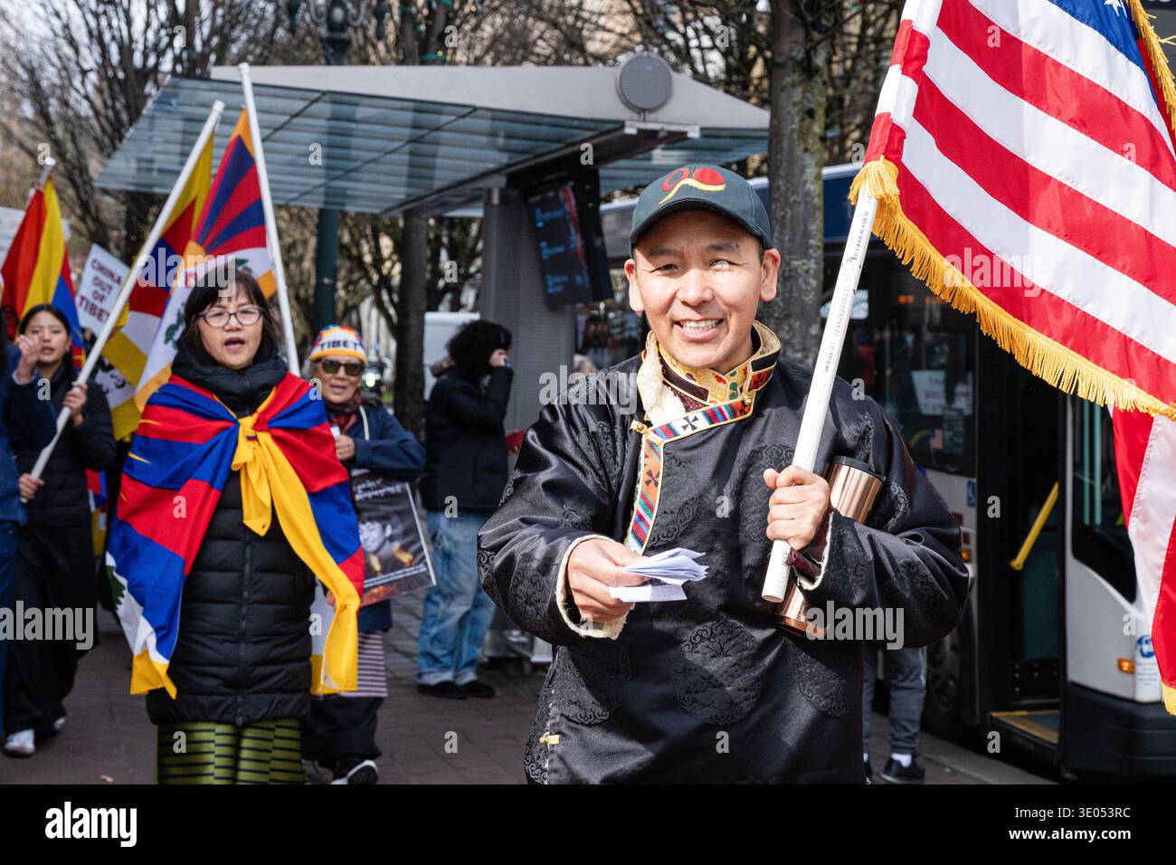 Portland, Oregon, Stati Uniti. 10 marzo 2026. Un uomo che indossa un tradizionale abito tibetano porta una bandiera degli Stati Uniti e tiene i materiali stampati mentre i partecipanti si riuniscono per una marcia del giorno della rivolta tibetana organizzata dalla Northwest Tibetan Cultural Association. Una donna drappeggiata in una bandiera tibetana cammina dietro di lui. Crediti: Norboe Photo/Alamy Live News Foto Stock