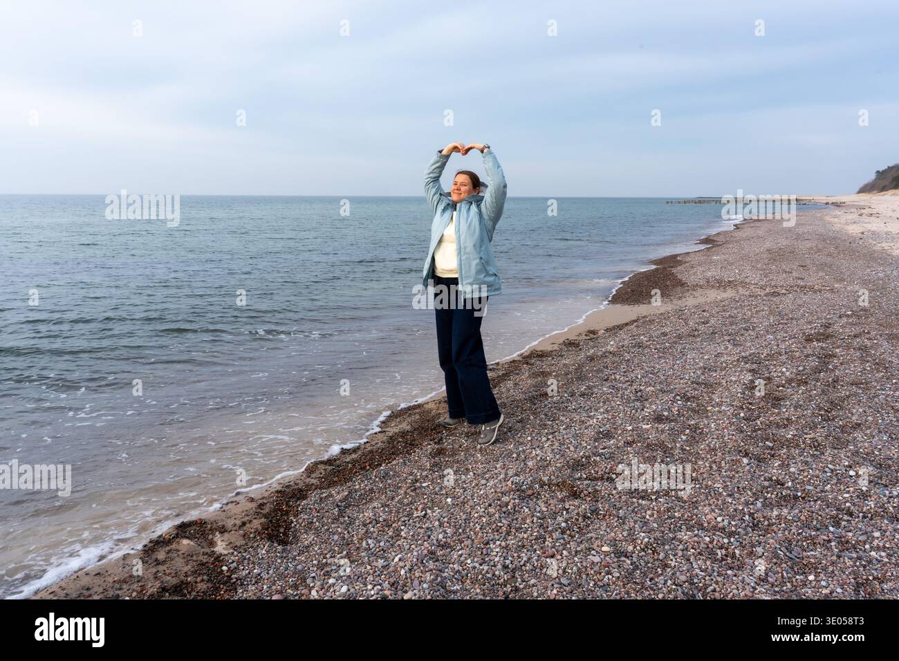Allegra giovane donna in giacca azzurra che dà forma al cuore con le mani sulla spiaggia rocciosa in una giornata fredda e nuvolosa. Foto Stock