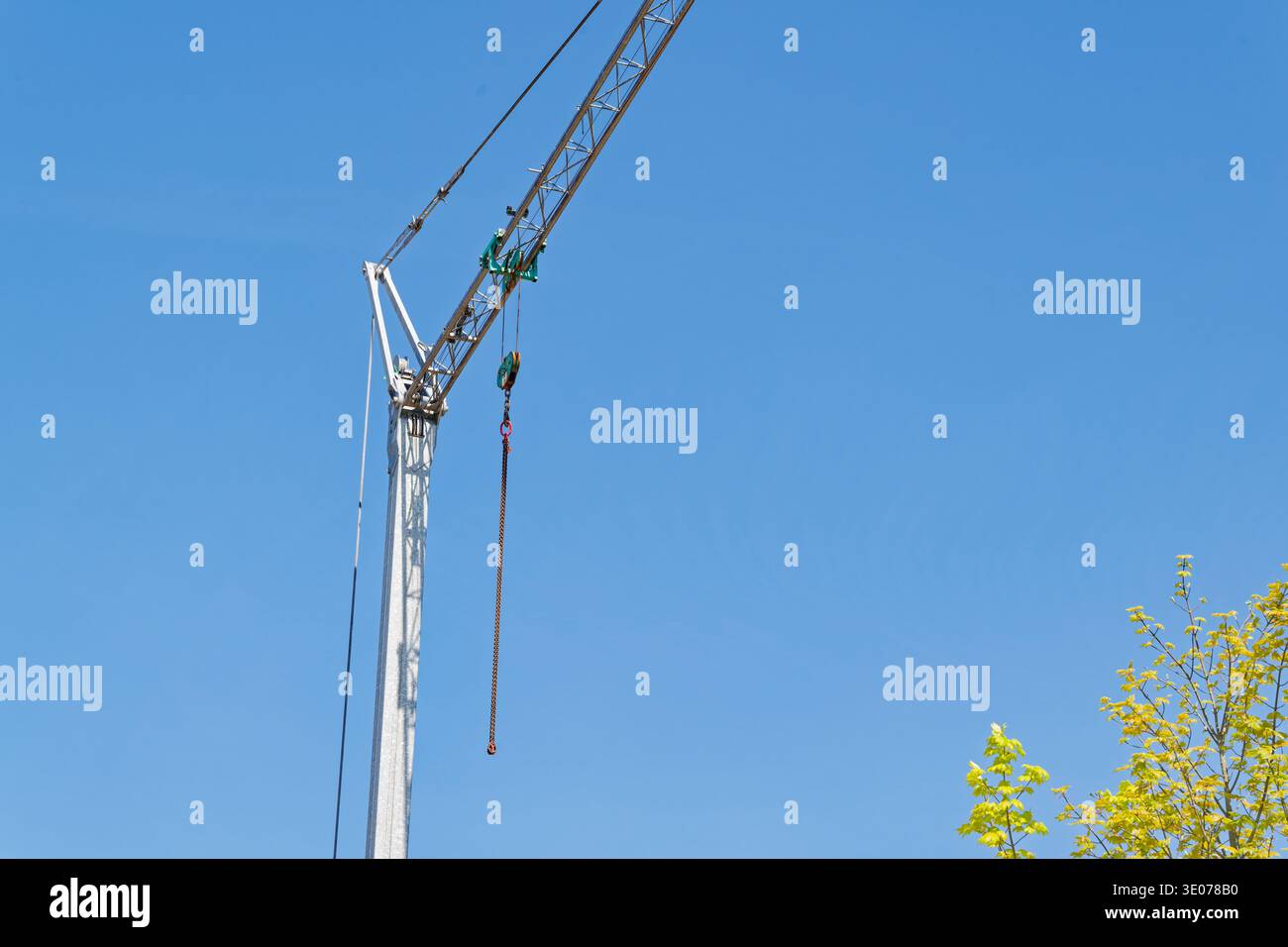 Un'alta gru da costruzione con un gancio sospeso e una catena si erge contro un cielo blu brillante con un albero nell'angolo, a simboleggiare l'edificio e lo sviluppo Foto Stock