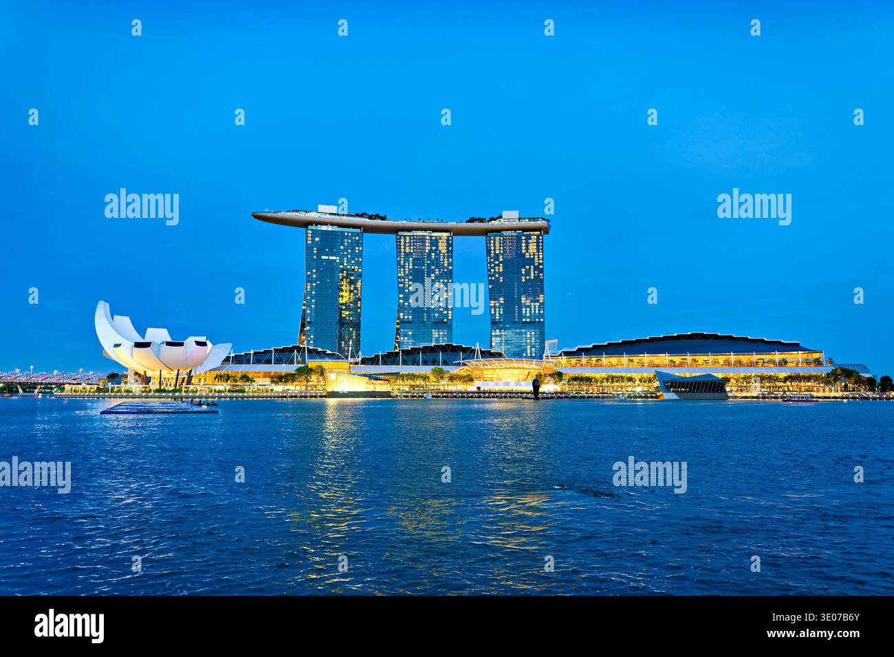 Le persone si godono la serata a Marina Bay mentre le luci della città iniziano a brillare e gli edifici riflettono sull'acqua durante il tramonto. Foto Stock