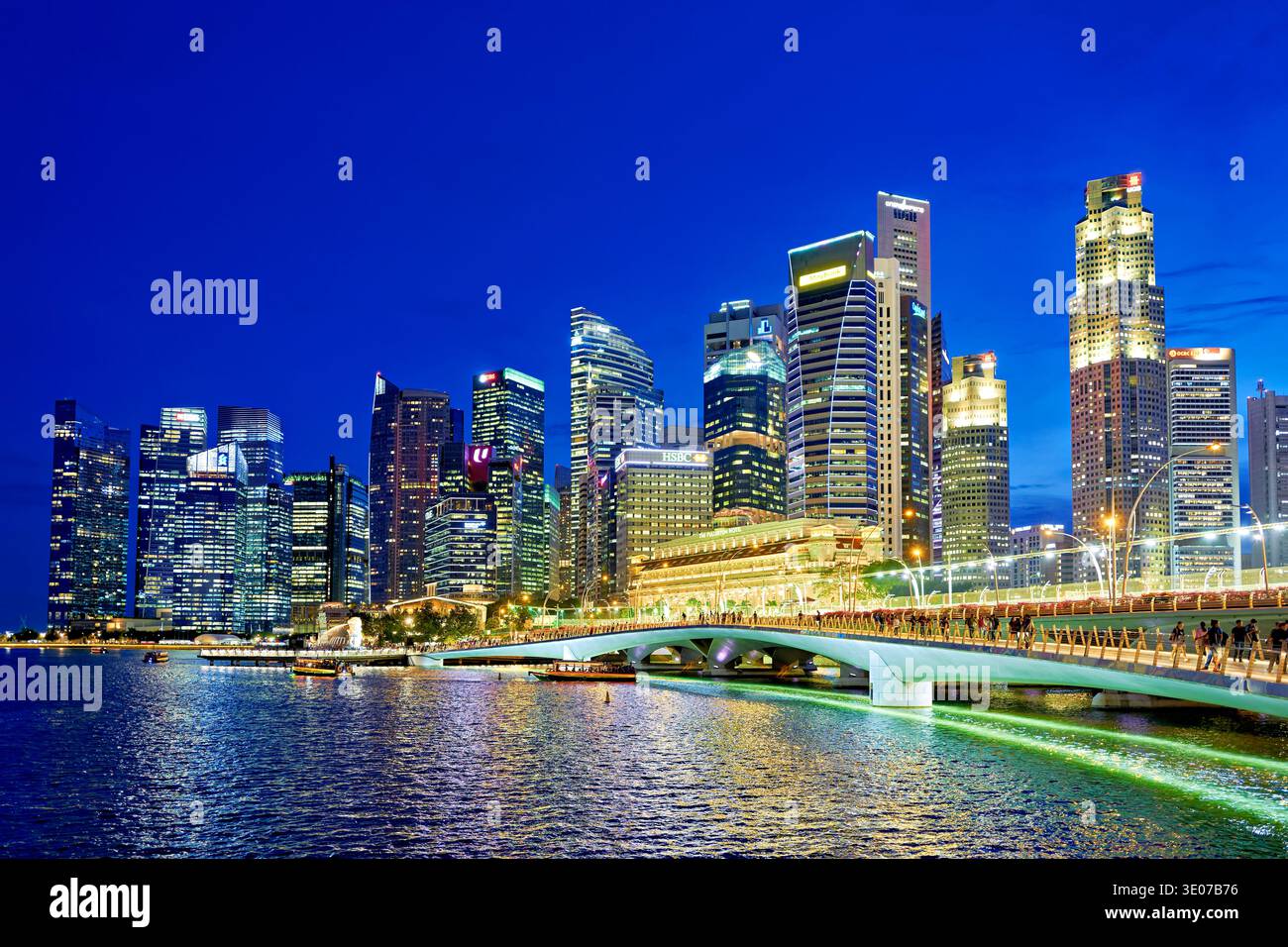 Le persone si godono la serata a Marina Bay mentre le luci della città brillano e riflettono sull'acqua. Lo skyline è mozzafiato sotto il cielo del tramonto. Foto Stock