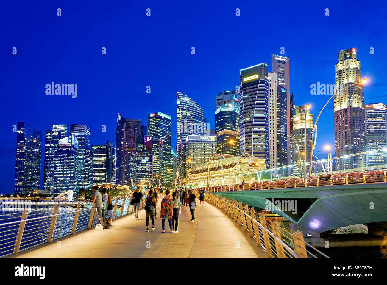 La gente cammina lungo il lungomare di Marina Bay mentre lo skyline della città si illumina durante il tramonto. Gli edifici riflettono sulla superficie dell'acqua. Foto Stock