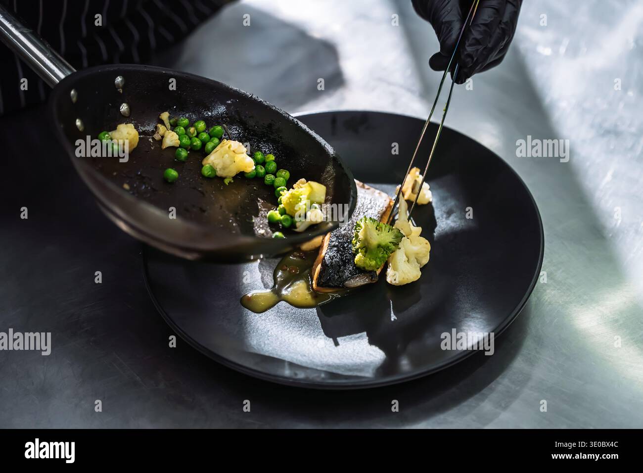 Primo piano di uno chef con guanti neri che prepara accuratamente verdure saltate su un piatto nero con pesce grigliato. Processo di placcatura culinaria di alta qualità in un p Foto Stock