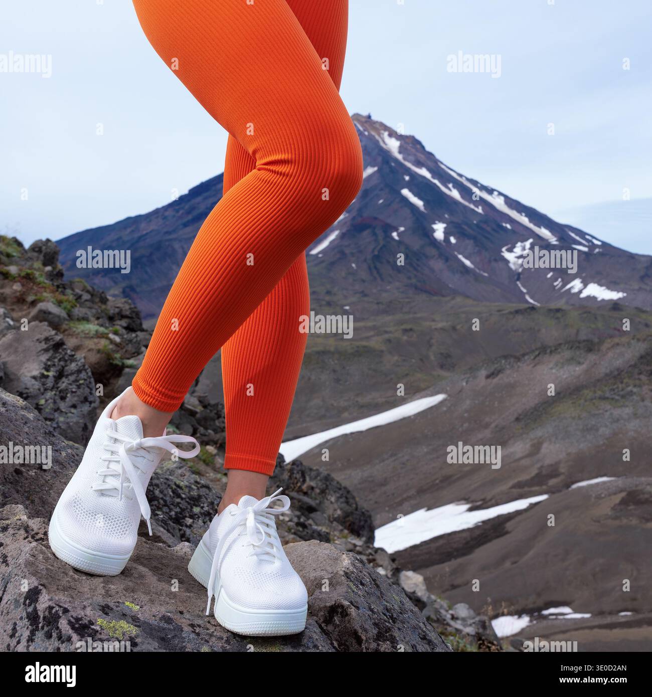 Primo piano delle gambe di una donna con leggings a coste arancione brillante e sneaker bianche. Si erge su un sentiero roccioso di montagna, con picco vulcanico nella ba Foto Stock