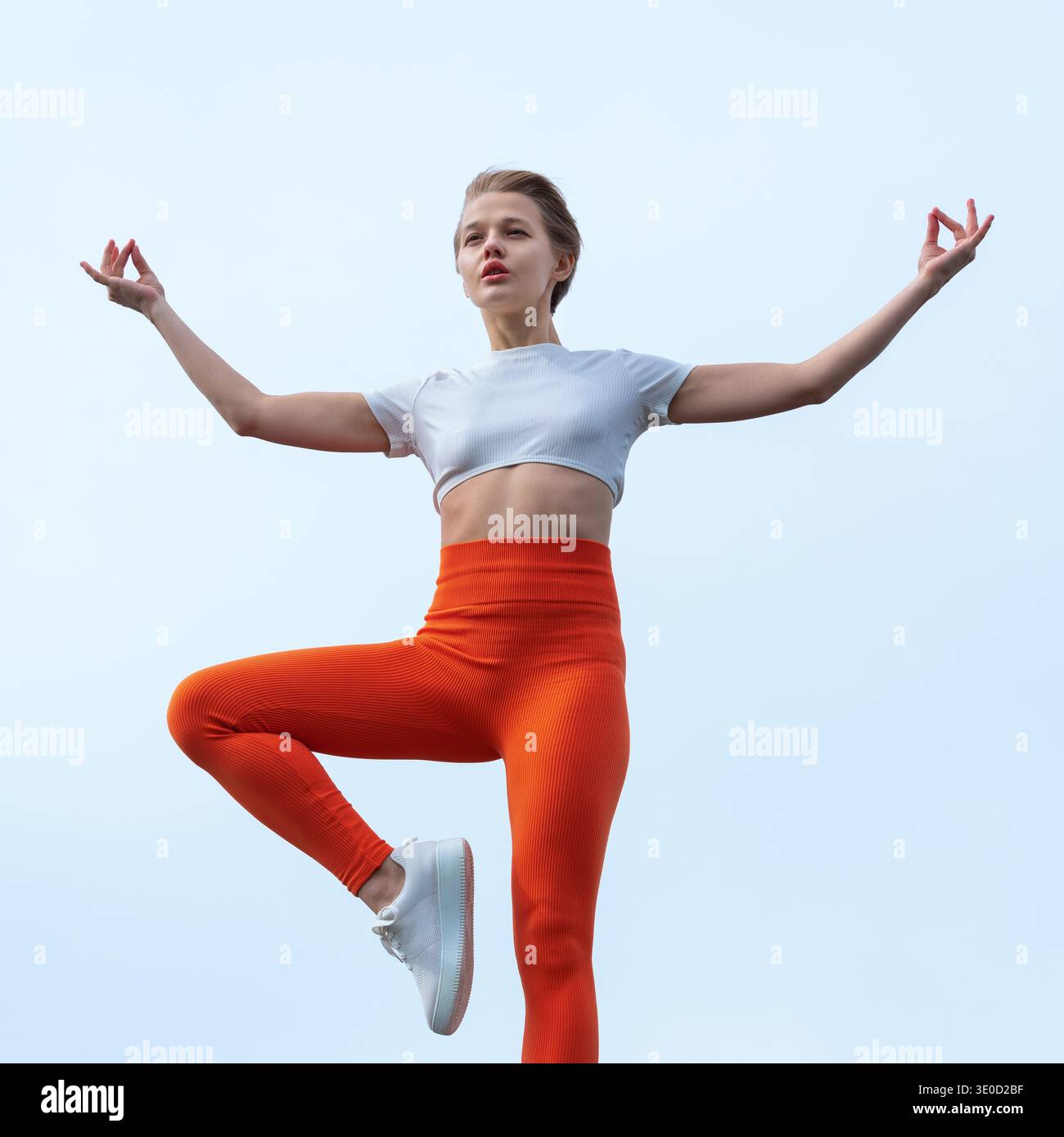 Una donna serena con leggings arancioni che fa una posa su un albero di yoga contro un cielo blu. La composizione minimalista e la luce soffusa esaltano il suo equilibrio, la sua messa a fuoco, una Foto Stock