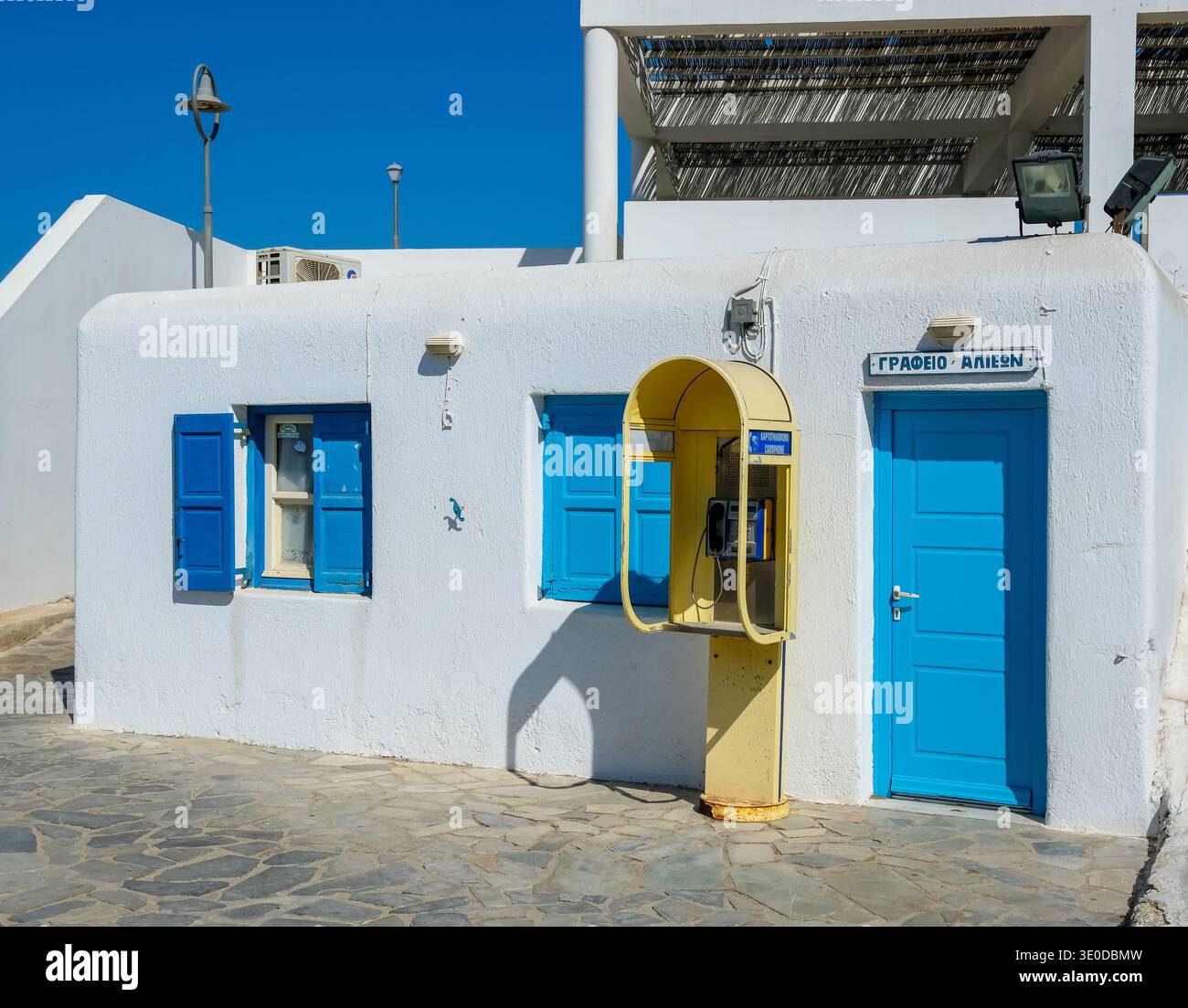 01.09.2025, Grecia, Mykonos, Mykonos - vecchia cassetta telefonica gialla di fronte a una casa bianca con persiane blu nelle strade della città vecchia di Mykonos. Foto Stock