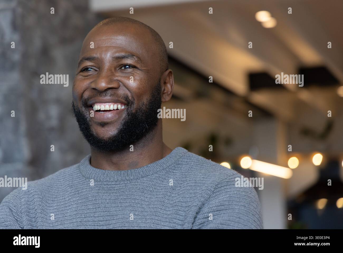 Uomo afro-americano sorridente, con girocollo in maglia grigia, in piedi vicino a una colonna di pietra in un moderno caffè Foto Stock