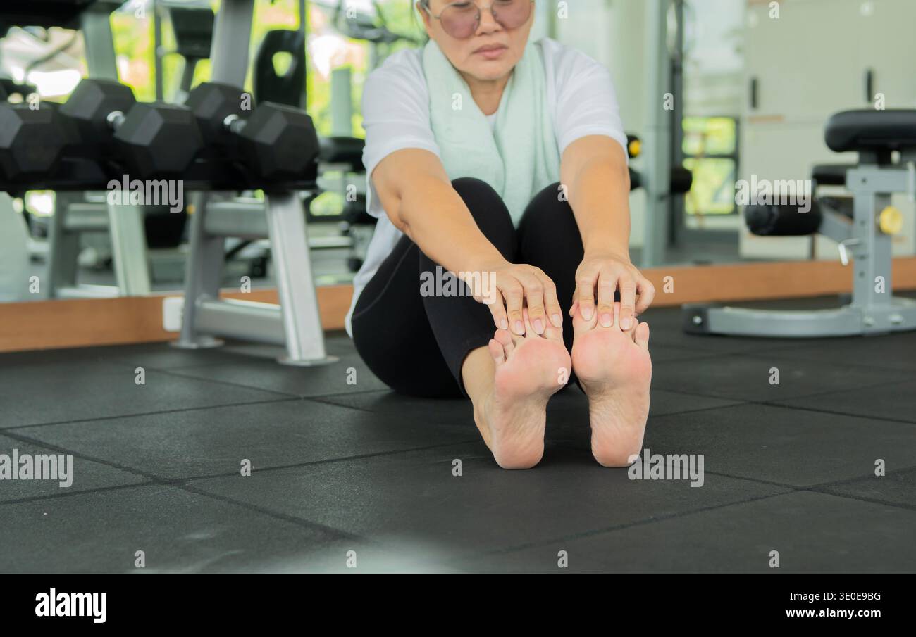 Crampi alle gambe. Donna anziana seduta sul pavimento e affetta da crampi alle gambe, dolore ai piedi in palestra. Concetto di problema di salute. Foto Stock