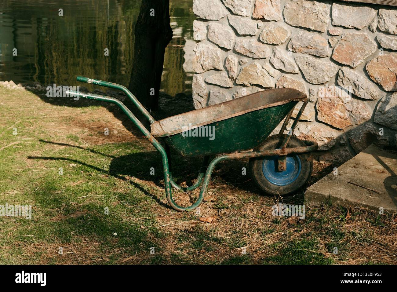 Carriola metallica verde rustica accanto a un muro di pietra vicino alle acque calme del lago. Concetto di attrezzi da lavoro rurali, attrezzature da giardinaggio e campagna Foto Stock