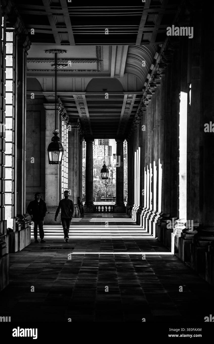 Due persone attraversano un corridoio della Greenwich University. La luce del sole crea ombre sul terreno e sulle pareti. L'architettura mostra il design dello spazio. Foto Stock