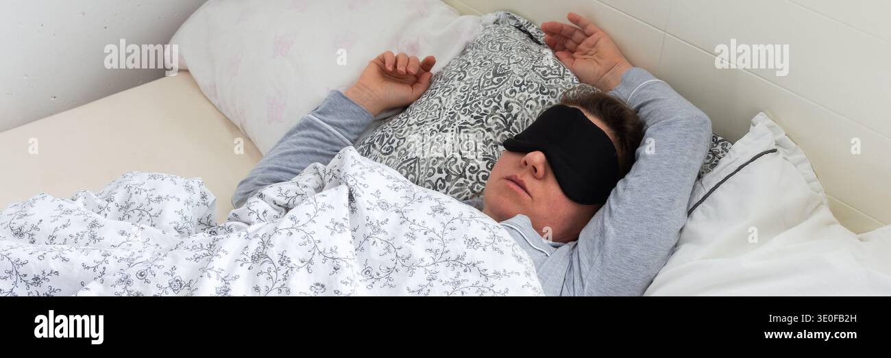 Una donna di 50 anni dorme nel suo letto e indossa una maschera. E' circondata da cuscini e coperte, creando un'atmosfera tranquilla. Sunli Foto Stock