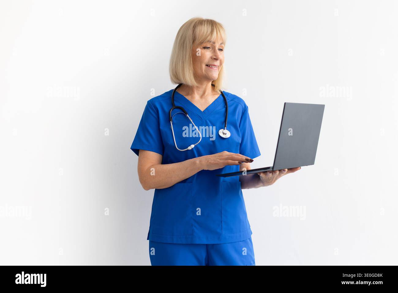 Professionista sanitario donna anziano che indossa scrub blu e stetoscopio che lavora su computer portatile Foto Stock