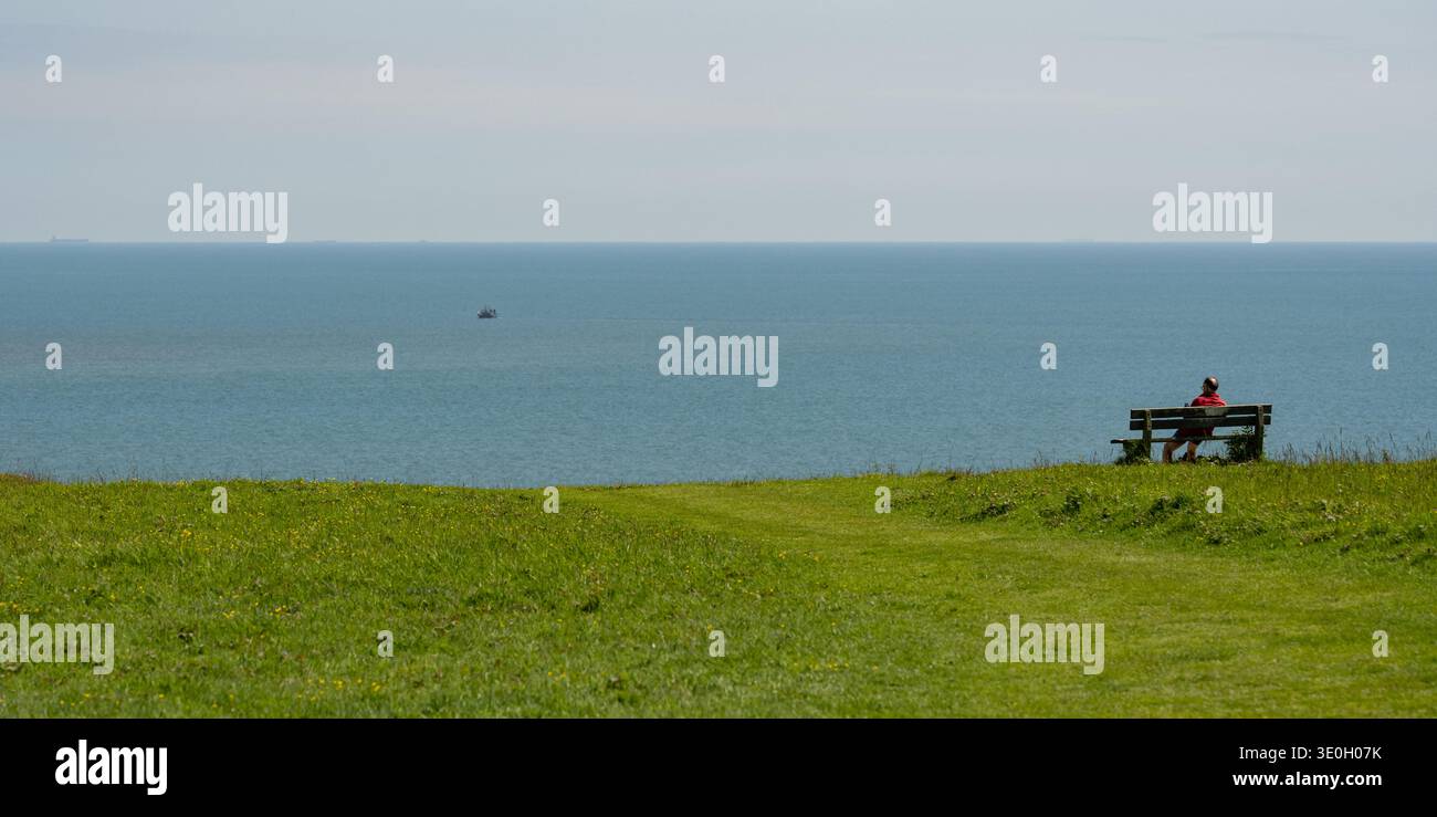 Uomo seduto su una panchina di legno a Seaford Head, East Sussex, affacciato sul Canale della Manica con un peschereccio in lontananza Foto Stock