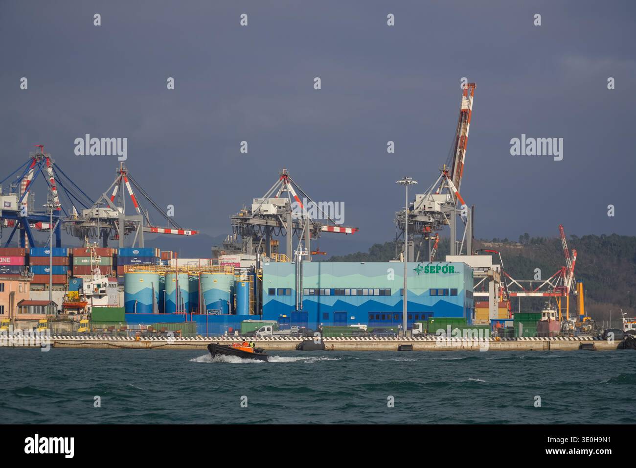 Porto di la Spezia, Italia. Struttura SEPOR visibile dietro le gru cargo e i container impilati sotto un cielo spettacolare. Piccole COSTOLE che accelerano sull'acqua. Foto Stock