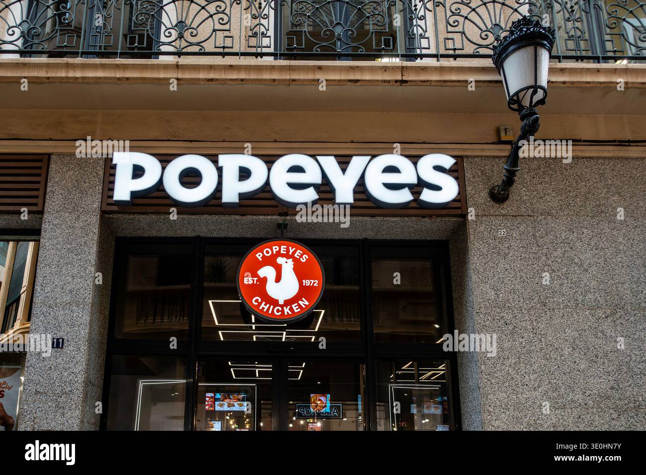 Vetrina illuminata di un ristorante Popeyes Chicken a Murcia, Spagna, con segnaletica moderna sotto un balcone con ringhiere in ferro ornate. Foto Stock