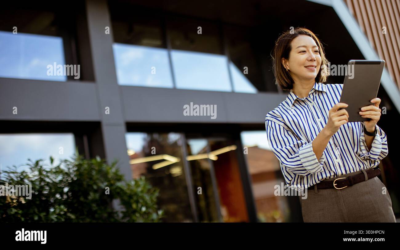 Una donna coreana sta fuori da un edificio moderno mentre usa un tablet. Sorride e sembra interagire con il dispositivo. L'impostazione è luminosa e. Foto Stock