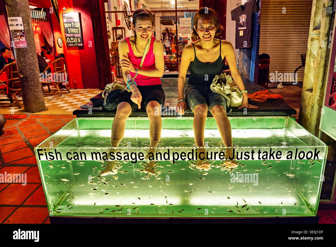 Due donne siedono sul bordo di una vasca, sorridendo mentre i pesci nuotano intorno ai loro piedi in una spa per piedi a Siem Reap, Cambogia. Foto Stock