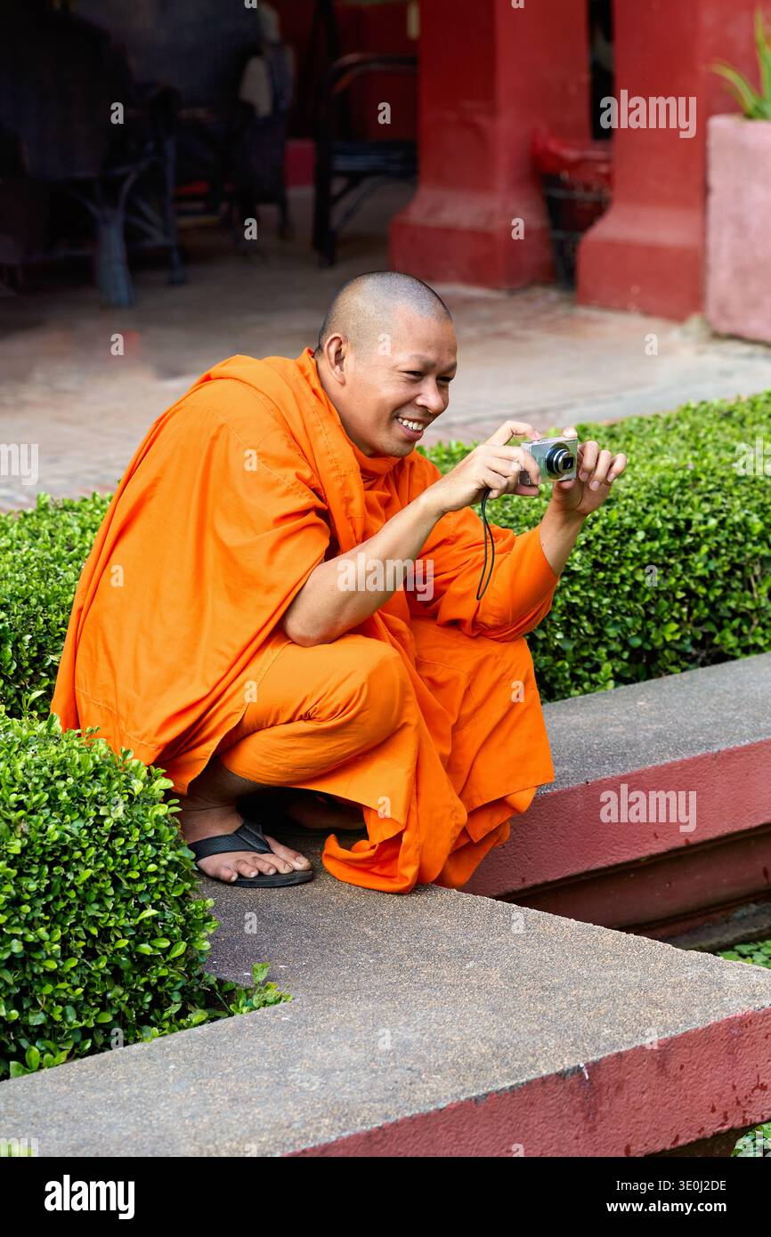 Un monaco in abiti arancioni accovacciato vicino ai cespugli, utilizzando una fotocamera digitale e sorridendo mentre cattura un momento a Phnom Penh. Foto Stock