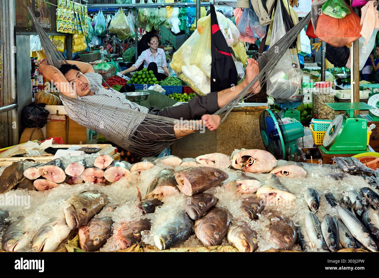 Un commerciante si rilassa in un'amaca sul suo banco di pesce mentre una donna vende verdure in un mercato cambogiano affollato. Foto Stock