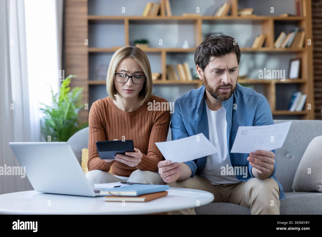 Coppia stressata che analizza le spese e le bollette finanziarie, utilizza una calcolatrice e un laptop per gestire il debito e il budget delle famiglie, preoccupata per la loro situazione economica Foto Stock