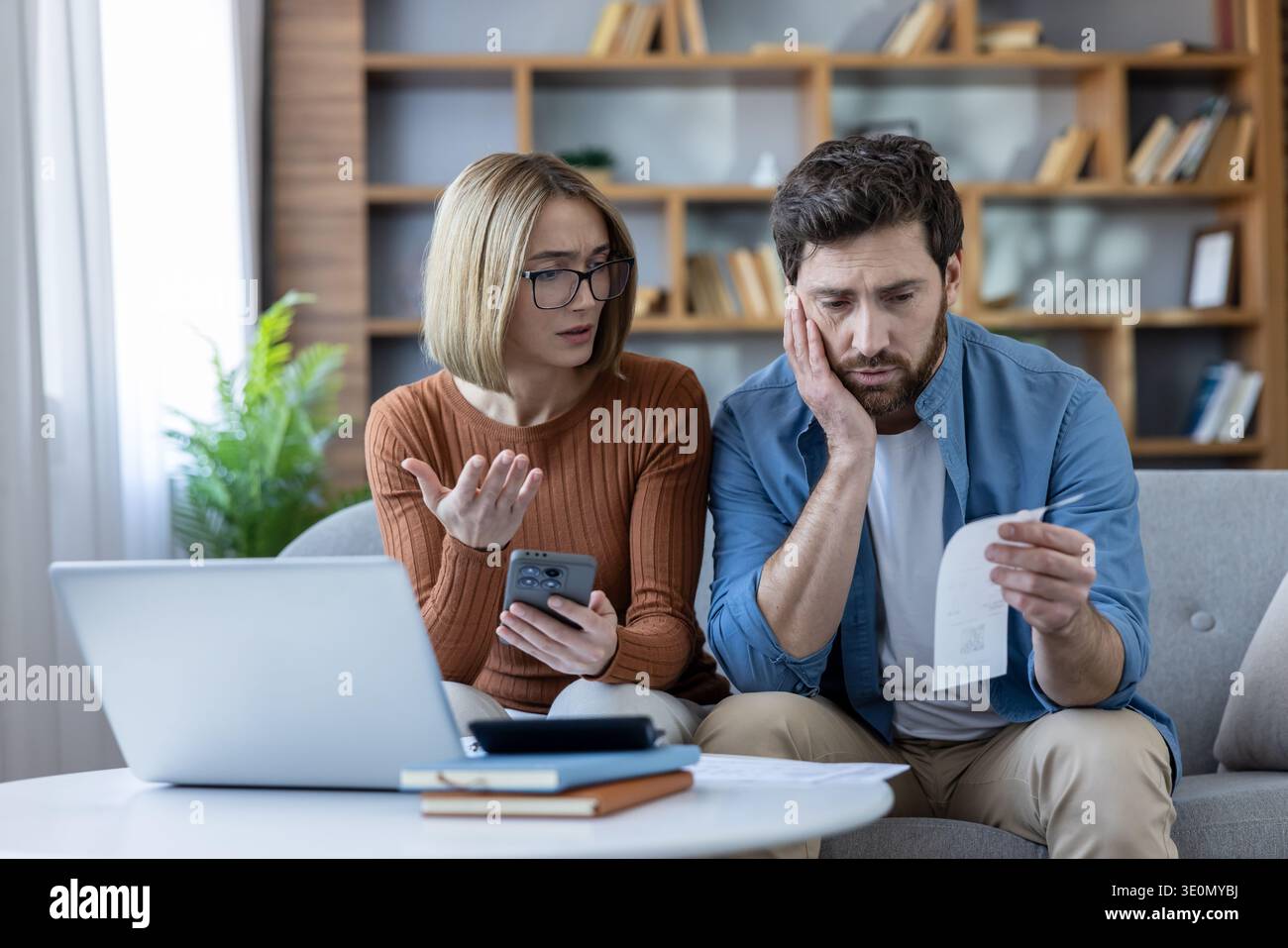 Coppia preoccupata su un divano di fronte a problemi finanziari e debiti, calcolando il budget della famiglia con un portatile e un telefono, esprimendo preoccupazione per l'aumento delle spese e la crisi economica Foto Stock
