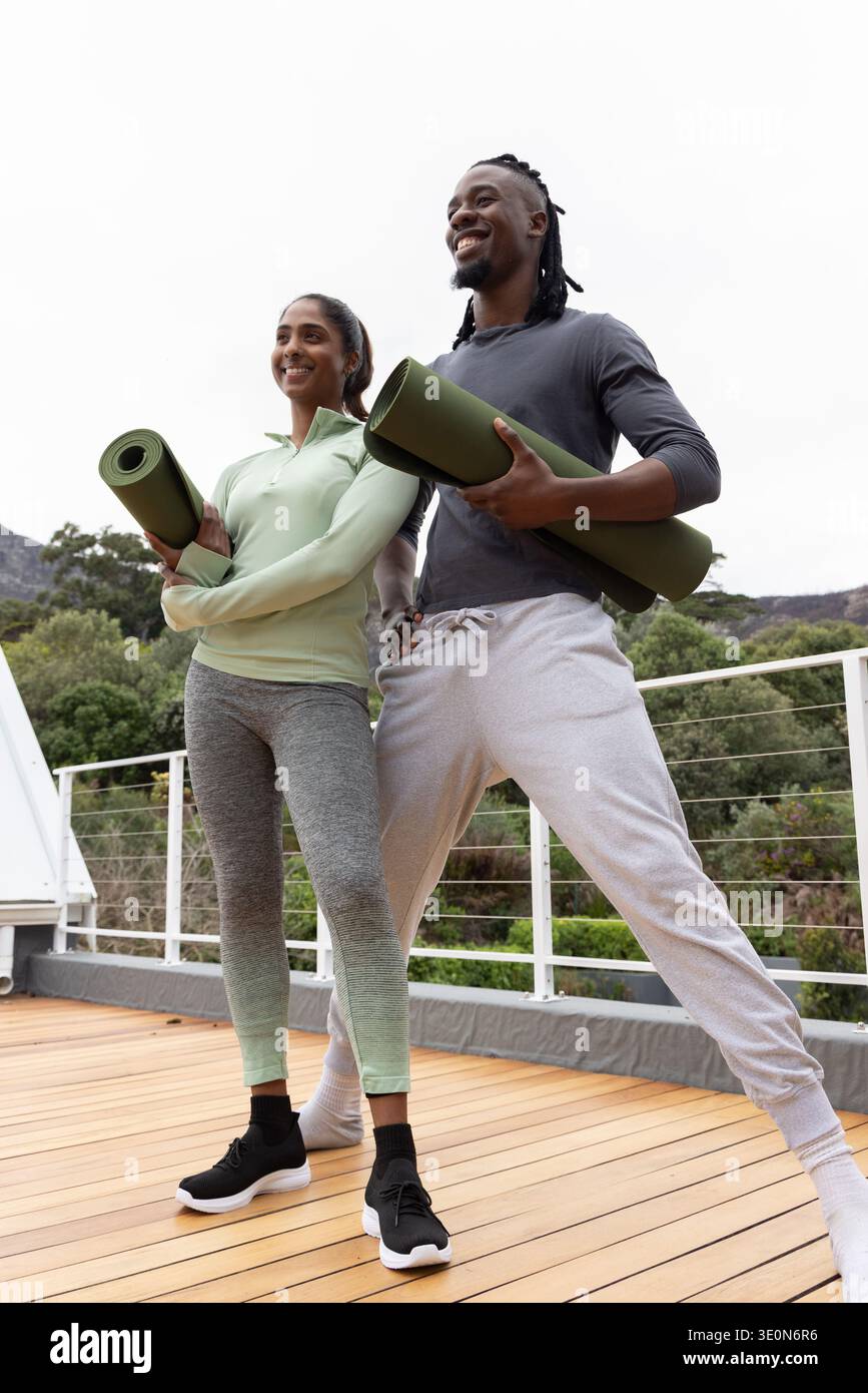 Amici diversi in piedi su un ponte di legno rialzato con tappetini da yoga verdi arrotolati, indossando abbigliamento sportivo Foto Stock