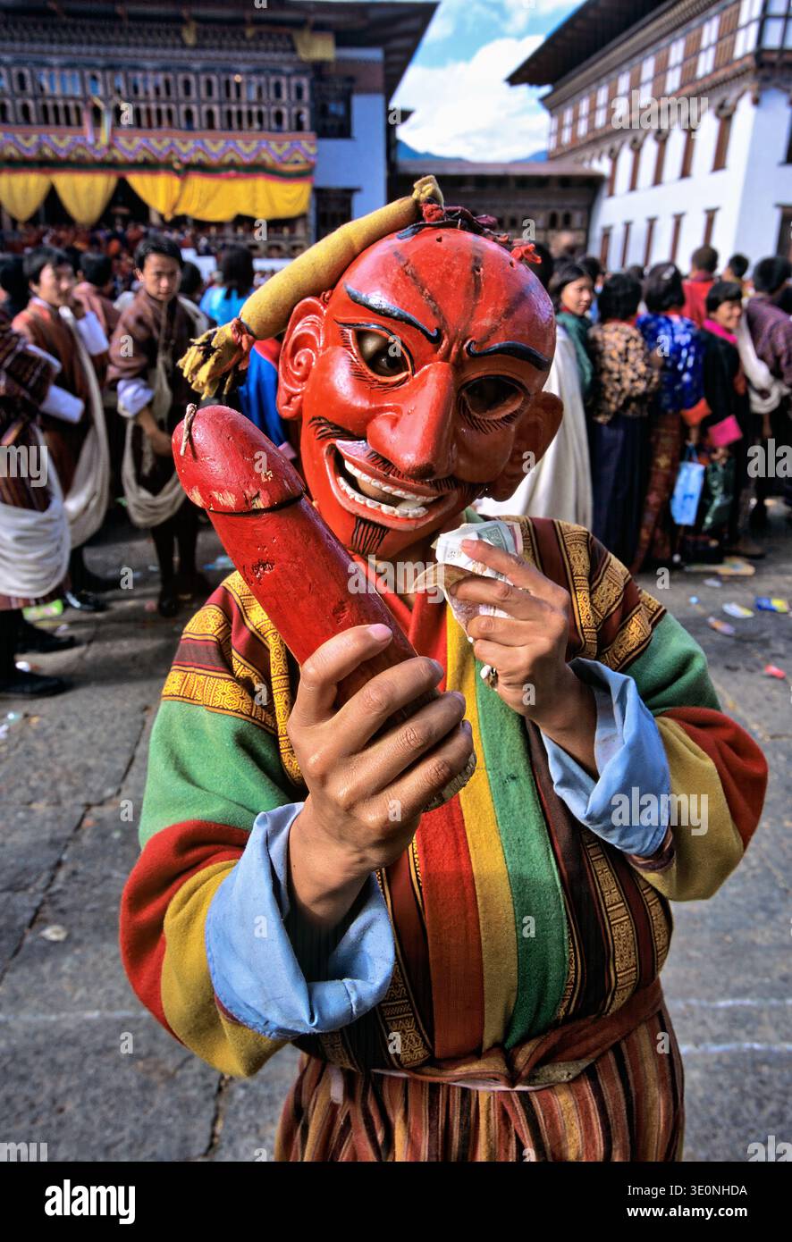 Una figura mascherata tiene in mano un fallo di legno e denaro durante una festa in Bhutan, che celebra la fertilità e protegge gli spiriti maligni. Foto Stock