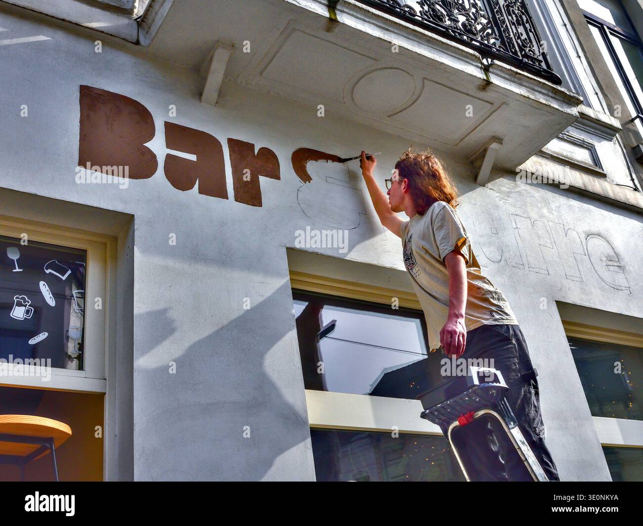 Giovane su una scala che dipinge la parola "Bar" sulla parete laterale di un bistrot - Saint-Gilles, Bruxelles, Belgio. Foto Stock