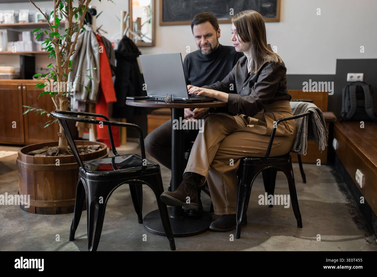 Uomo e donna professionisti che discutono di progetti e utilizzano computer portatili su una panca in legno in ufficio con decorazioni ad albero. Discussione di squadra, pianificazione, notebook, decorazione di alberi Foto Stock