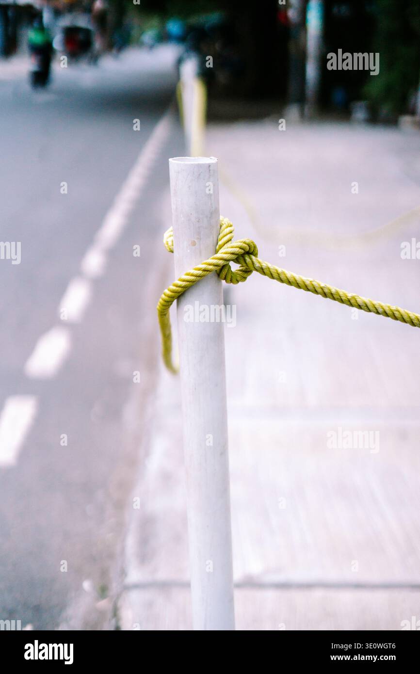 Una corda giallo-verde brillante strettamente legata con un nodo intorno a un palo bianco, adagiata su una strada urbana leggermente sfocata con un marciapiede e una strada in d Foto Stock
