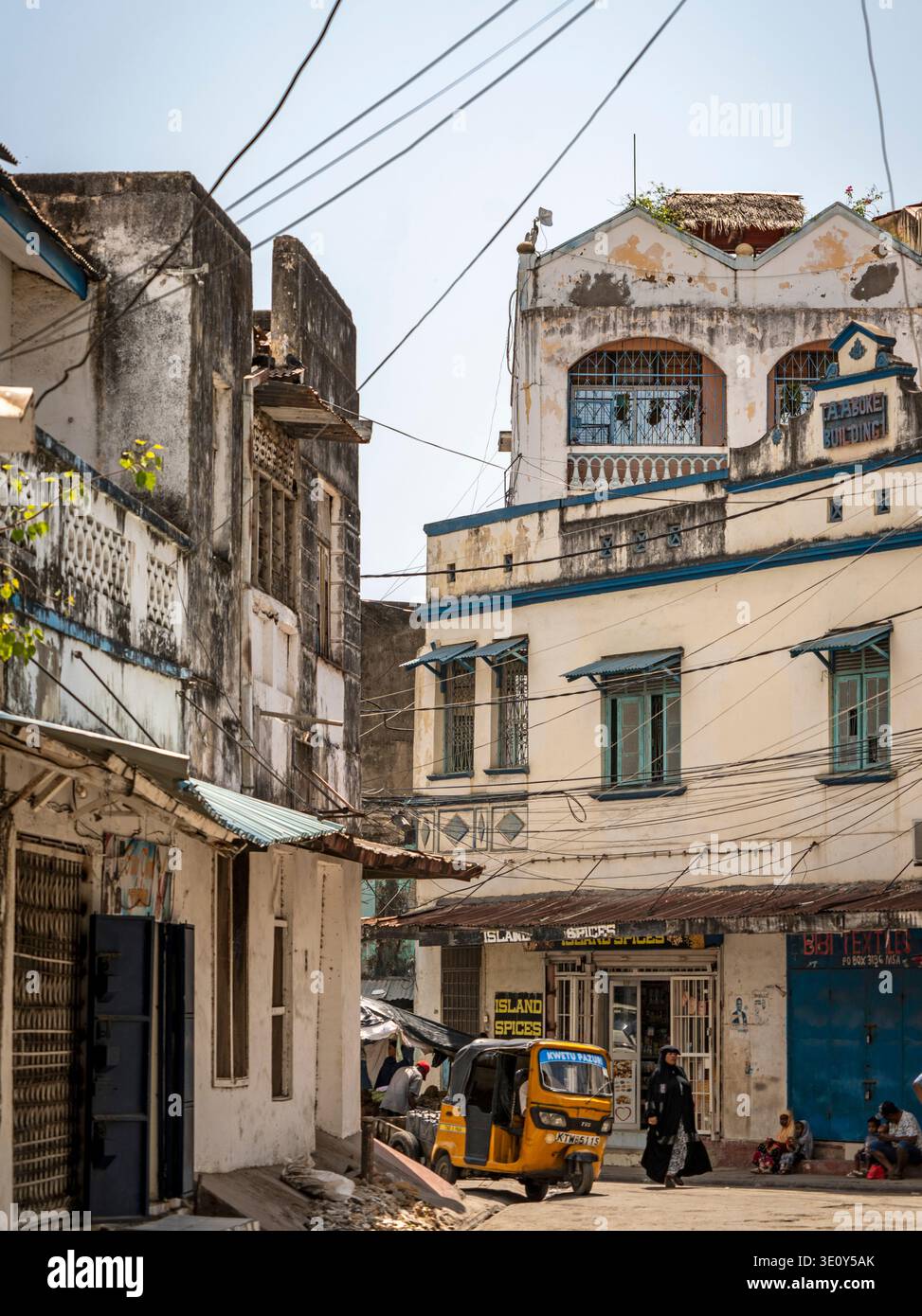Scena di strada in un'antica città costiera con edifici intemprati, linee elettriche aggrovigliate, piccoli negozi, pedoni e un tuk-tuk giallo sotto il sole Foto Stock