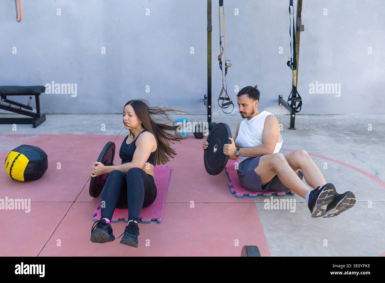 Due atleti stanno eseguendo torsioni russe con piastre di peso, rafforzando i loro muscoli muscolari in un ambiente di palestra all'aperto, circondato da vari fitne Foto Stock