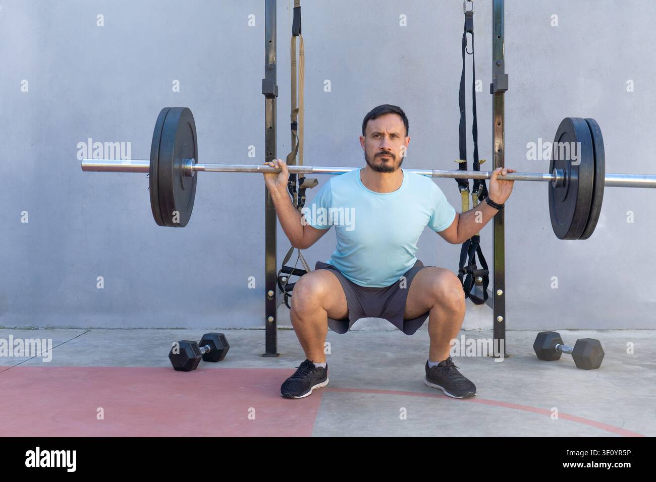 Atleta maschile concentrato che esegue squat da barbell in una palestra all'aperto, dimostrando forza e impegno per il fitness e abbracciando al contempo una forma attiva e curativa Foto Stock