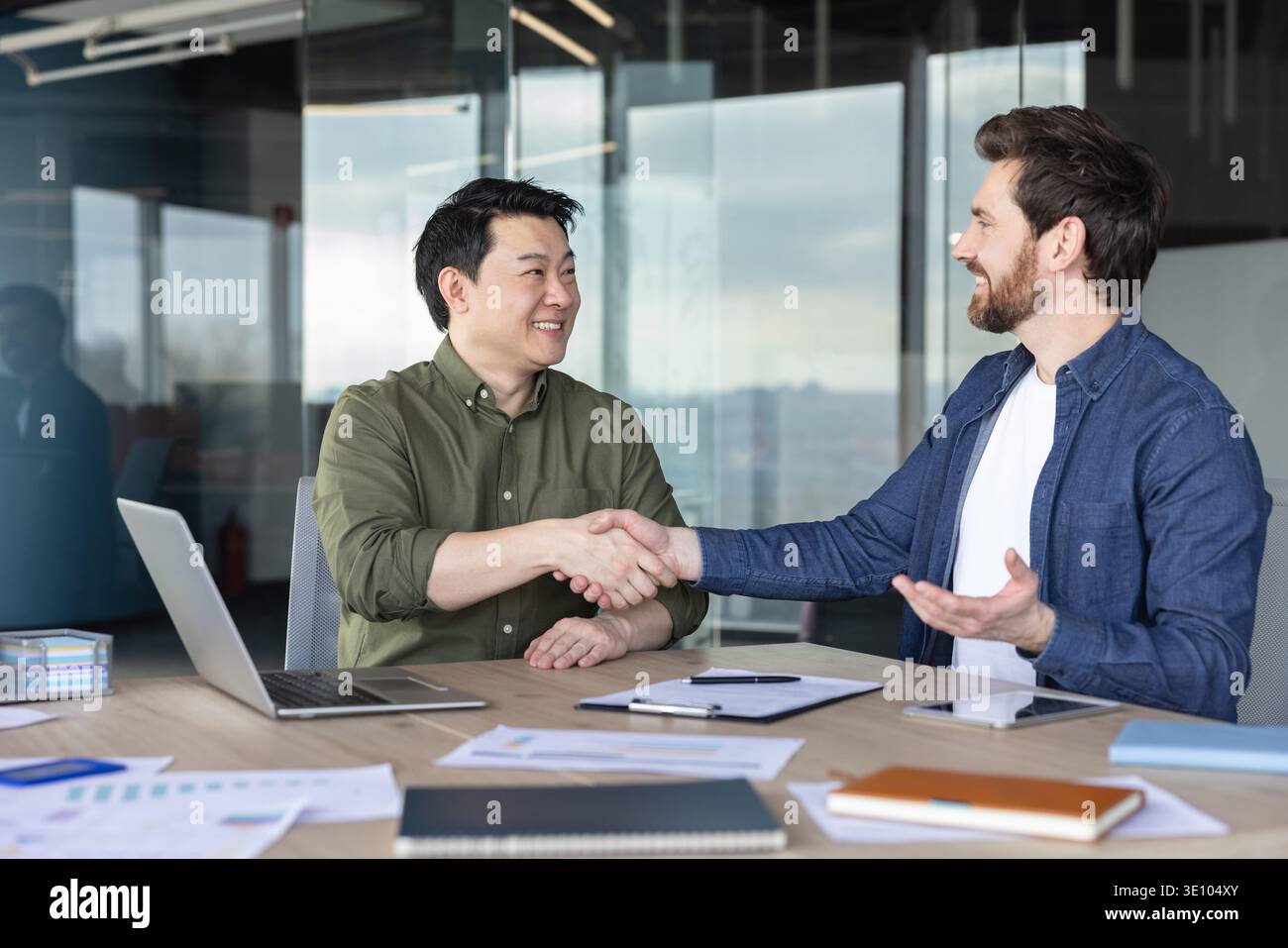 Due partner commerciali diversi stringono la mano, finalizzano un accordo e mostrano partnership, collaborazione e fiducia in un ambiente moderno per riunioni aziendali Foto Stock