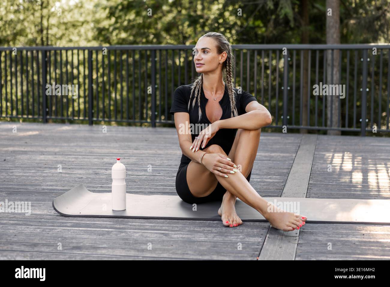 Attraente modello di donna atletica seduta su un tappetino da yoga nel parco verde della foresta, con bottiglia d'acqua dopo l'allenamento, allenamento fitness. Stile di vita sano, outdoo Foto Stock