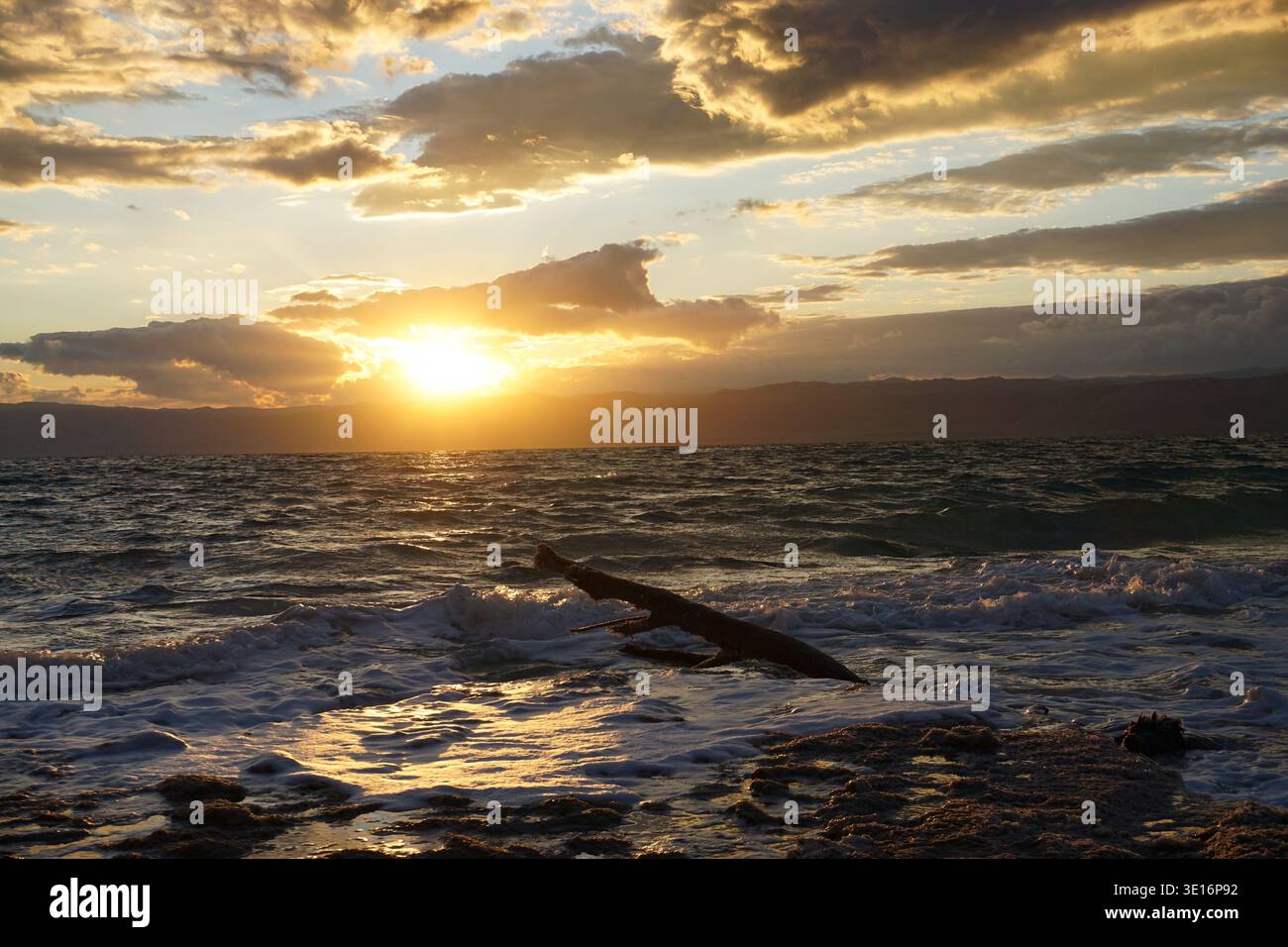 Tramonto sul Mar morto con le onde che raggiungono la costa rocciosa e la deriva in primo piano, la Giordania. Foto Stock