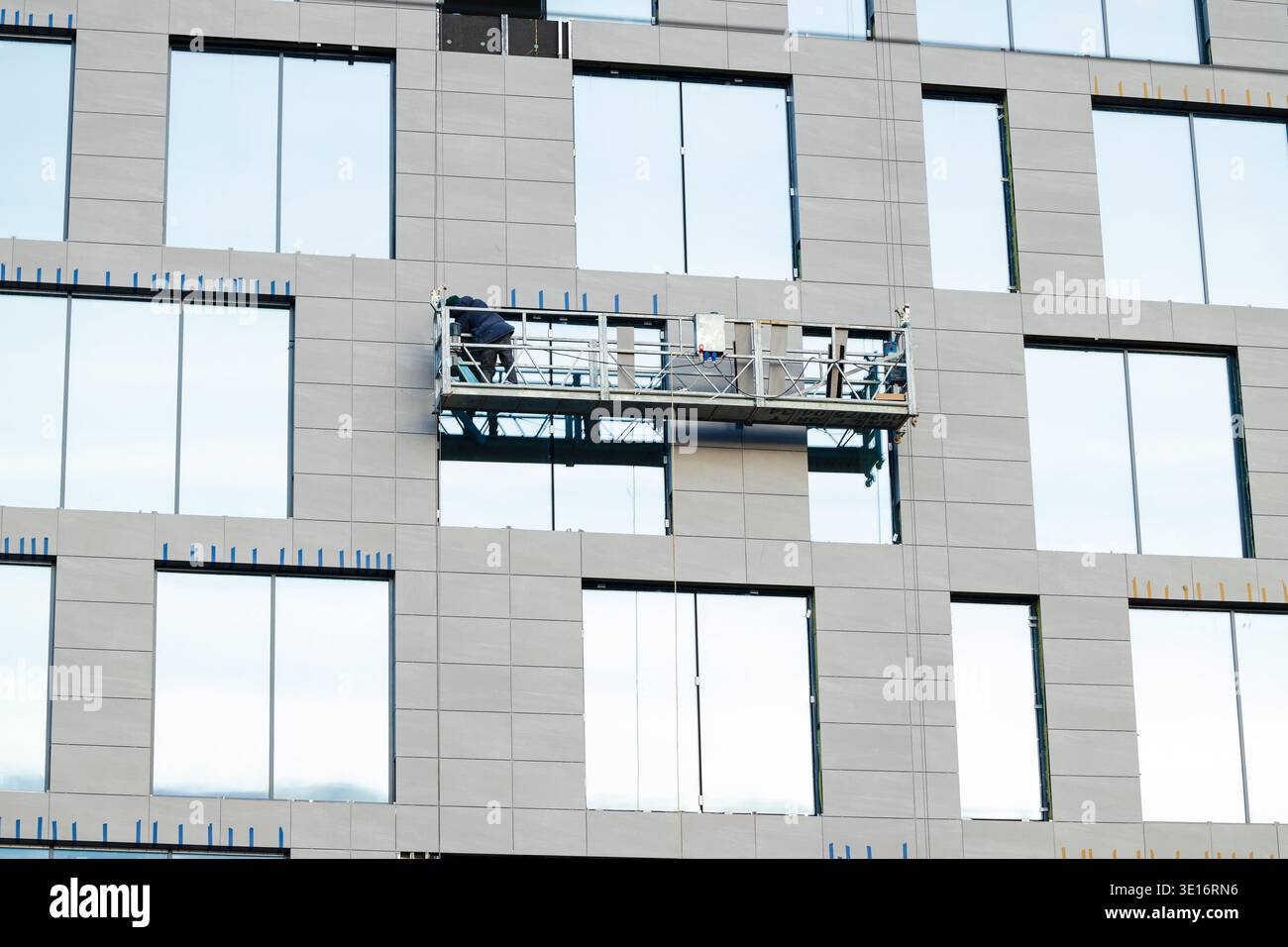 I lavoratori edili sono attivamente impegnati nell'installazione di pannelli esterni su un alto edificio utilizzando una piattaforma sospesa, che mostra lo sviluppo urbano Foto Stock