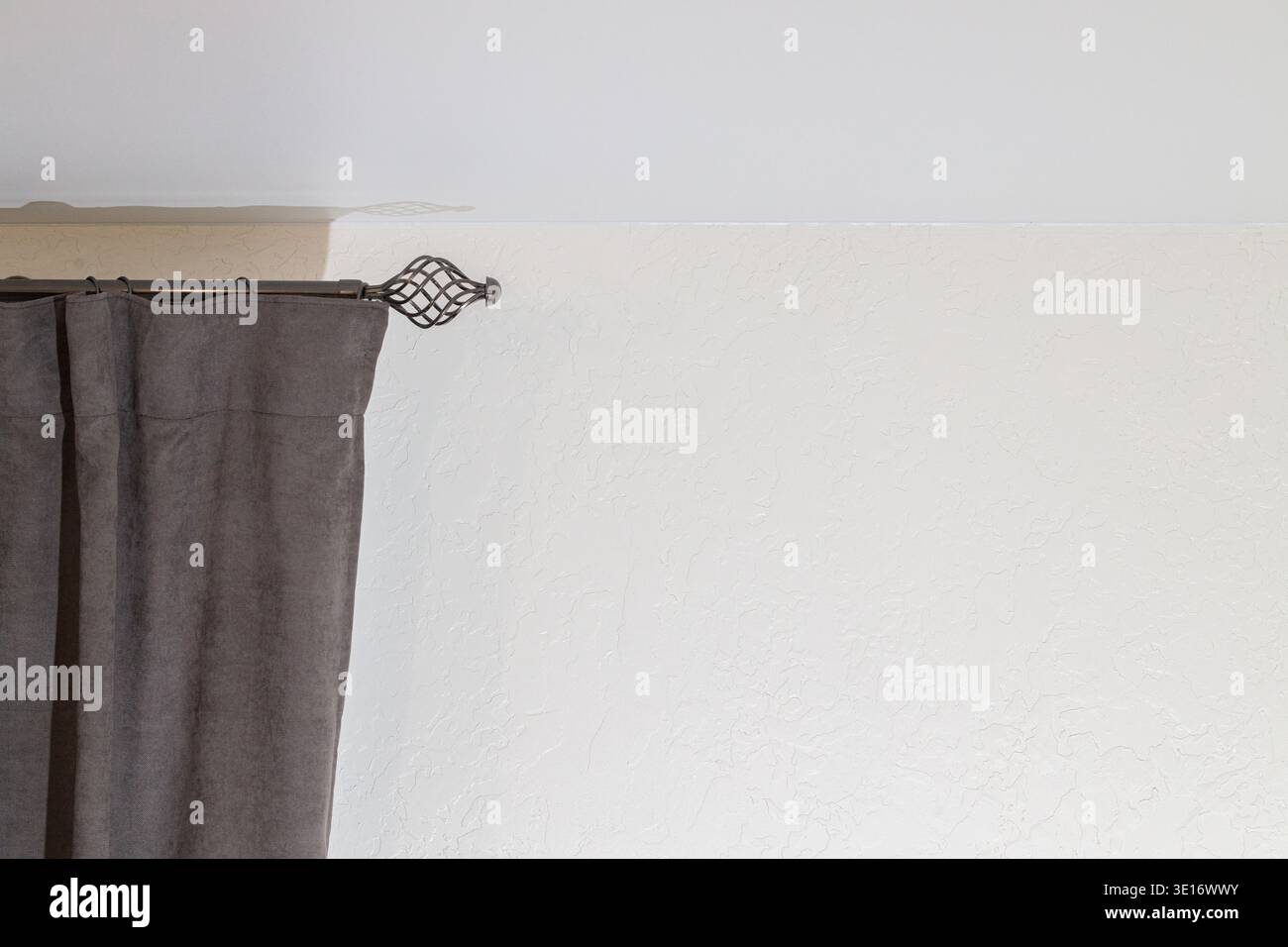 Una vista dettagliata di una tenda in tessuto grigio scuro parzialmente disegnata su un'asta decorativa in metallo, appoggiata su una parete testurizzata bianca Foto Stock