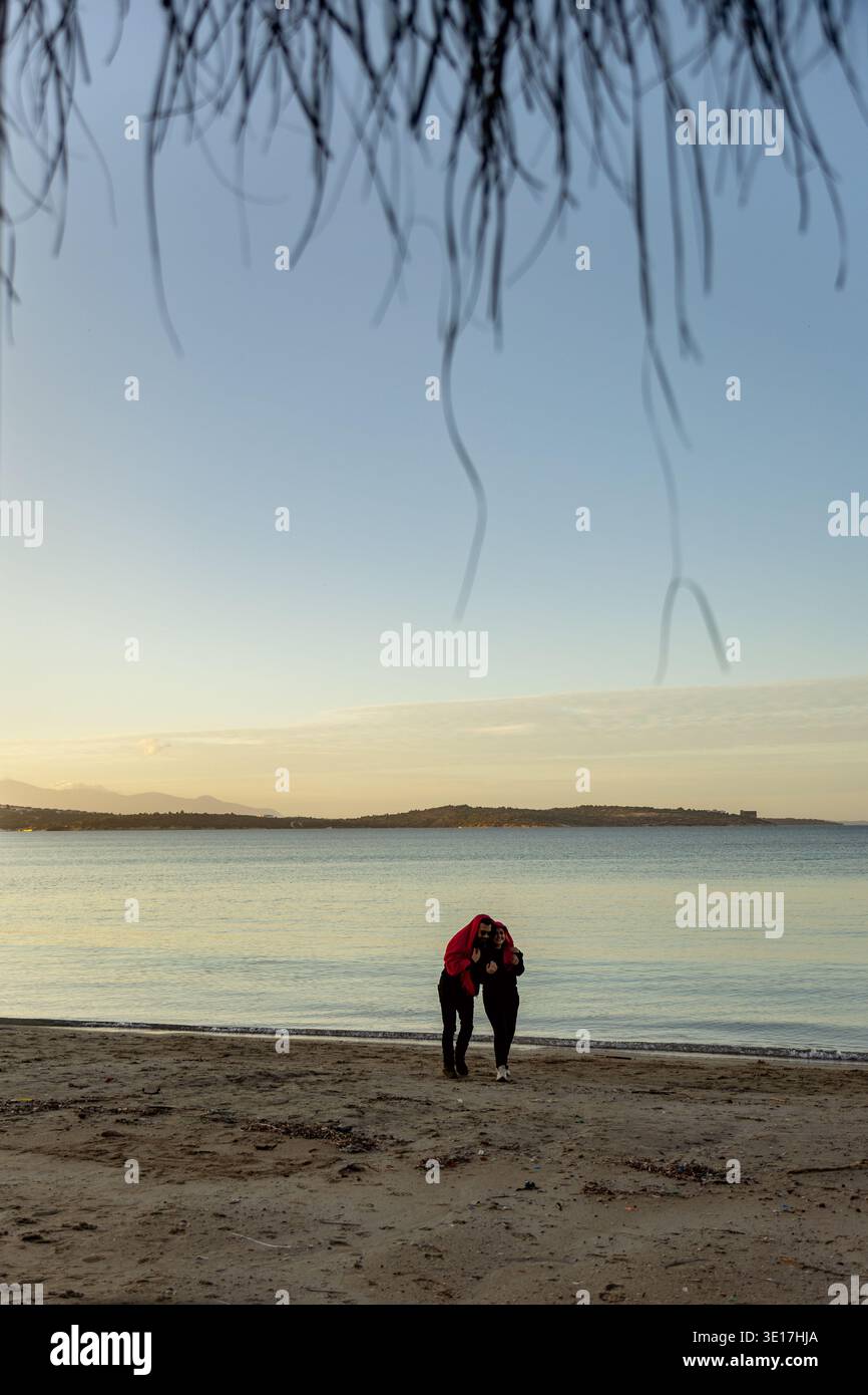 Una coppia gode di un momento sereno camminando lungo una spiaggia sabbiosa, avvolta tra le braccia dell'altra sotto un cielo limpido. Il mare si estende tranquillamente sul retro Foto Stock