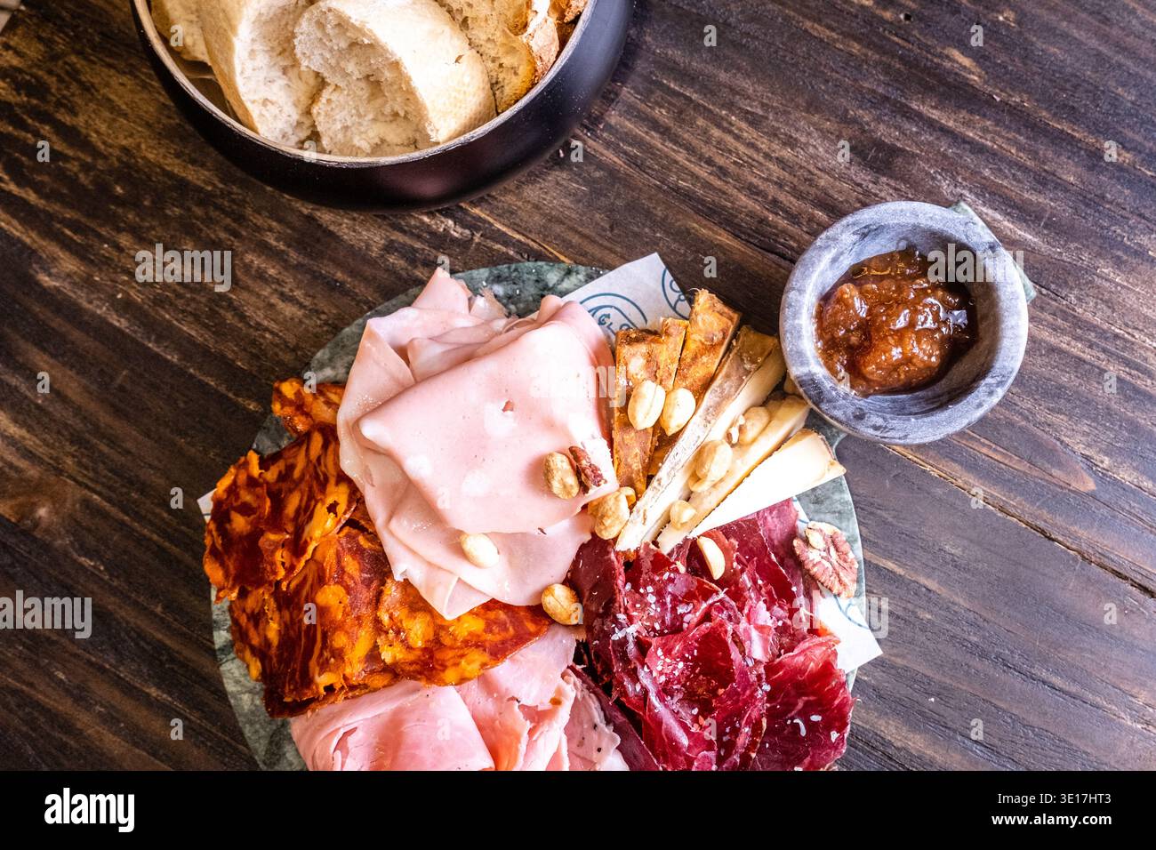 Vista dall'alto su un tagliere di salumi con una varietà di prosciutti stagionati, baguette affettate, cracker e una piccola ciotola di chutney, disposta ad arte Foto Stock