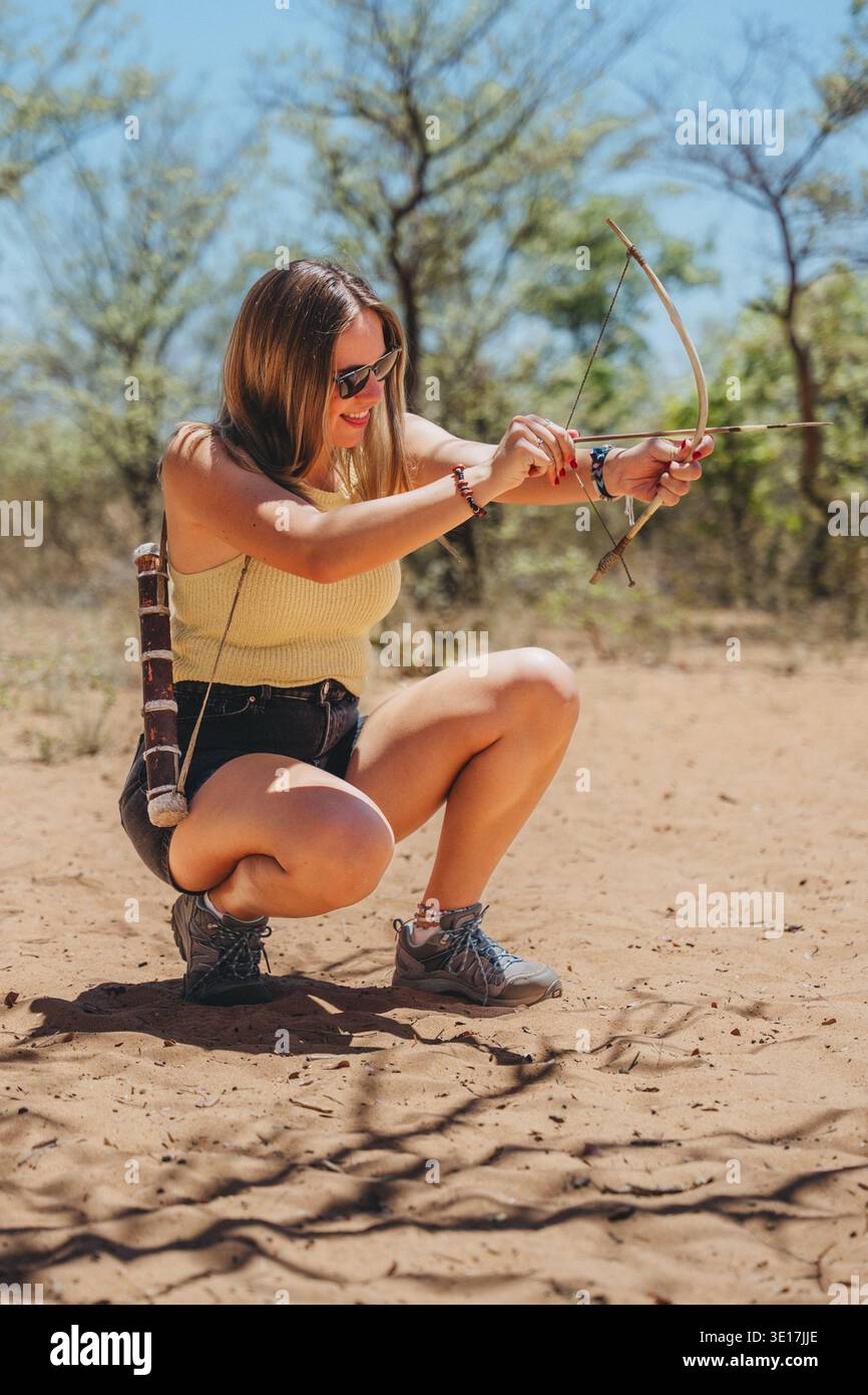 Si vede una donna che esplora il paesaggio namibiano, accovacciata su un terreno sabbioso mentre si prepara abilmente a sparare un arco tradizionale e una freccia sotto clea Foto Stock